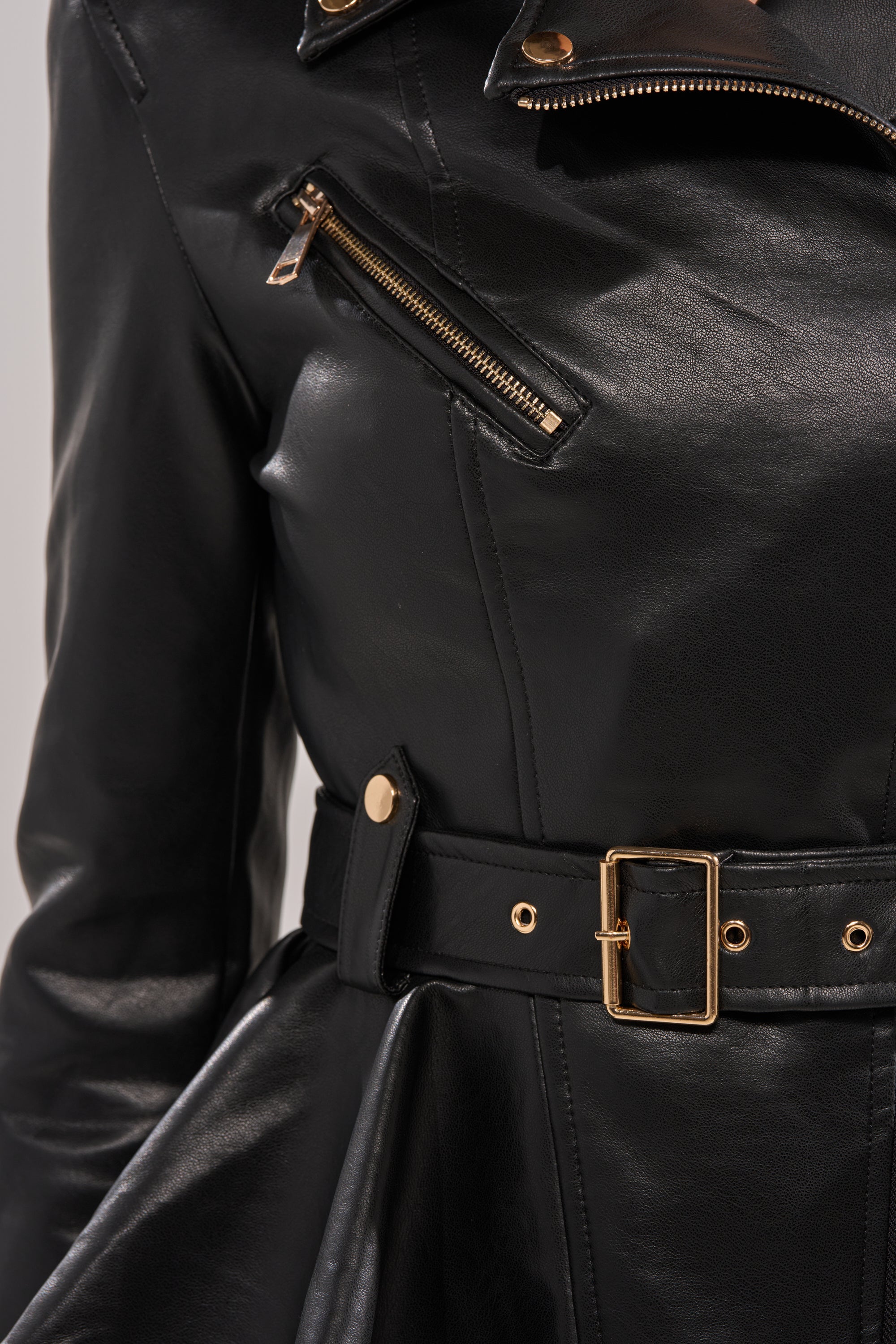 Close-up of the JULES PEPLUM MOTO jacket in black faux leather, featuring a gold chest zipper and matching gold-buckled waist belt, showcasing its shiny texture and stylish metal details. 8