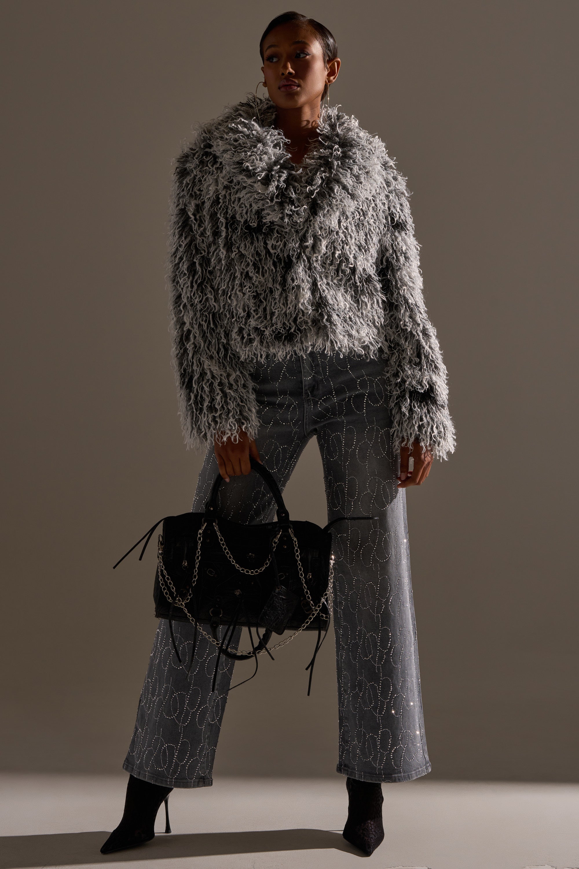 Wearing the RENDEZVOUS SHAGGY FAUX FUR coat, high-waisted printed jeans, black pointed boots, and holding a black chain-detailed handbag, a model poses against a neutral background.