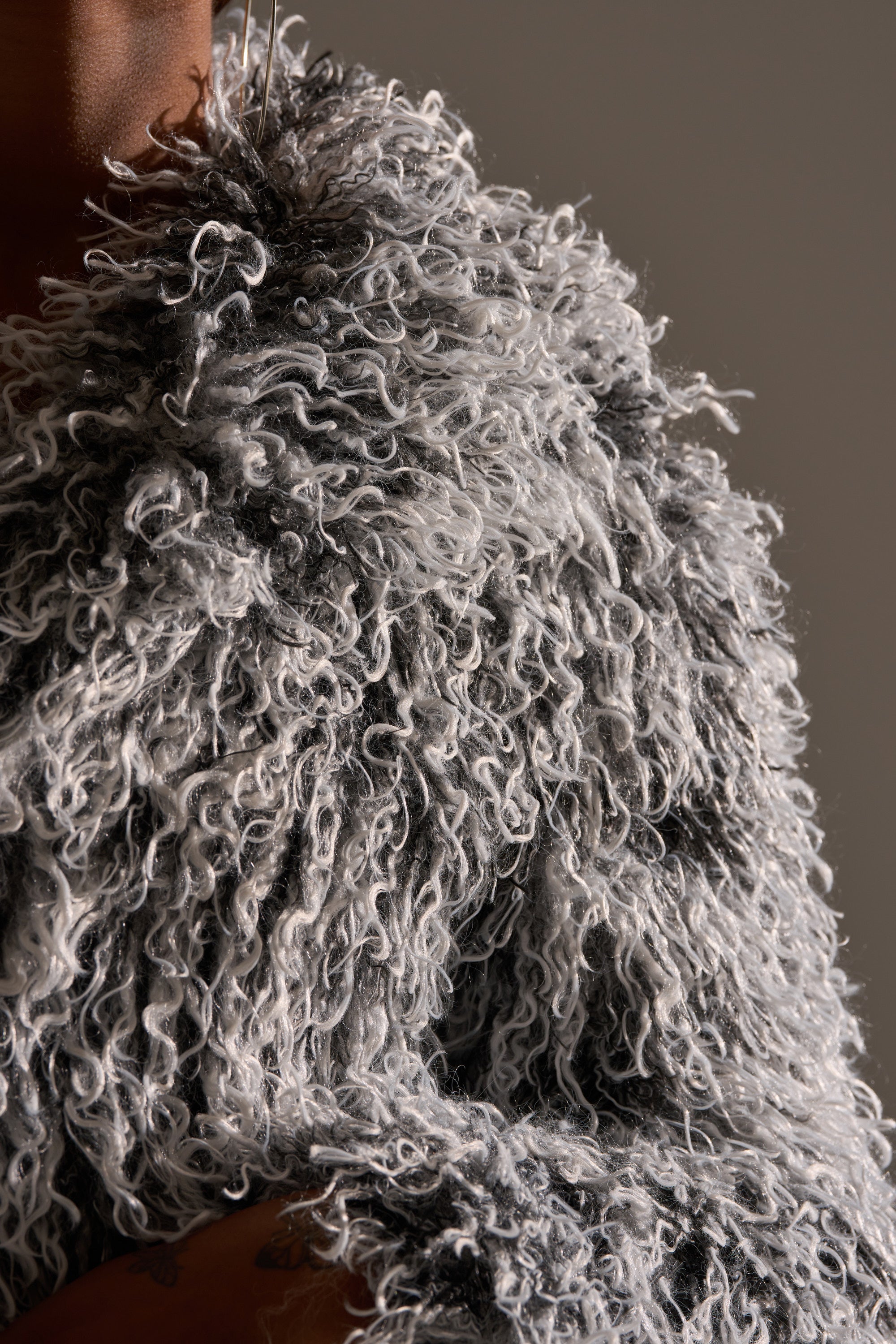 Close-up of a person wearing the RENDEZVOUS SHAGGY FAUX FUR coat, showing its textured, poodle-like finish, with only part of their face and hand visible against a neutral background.
