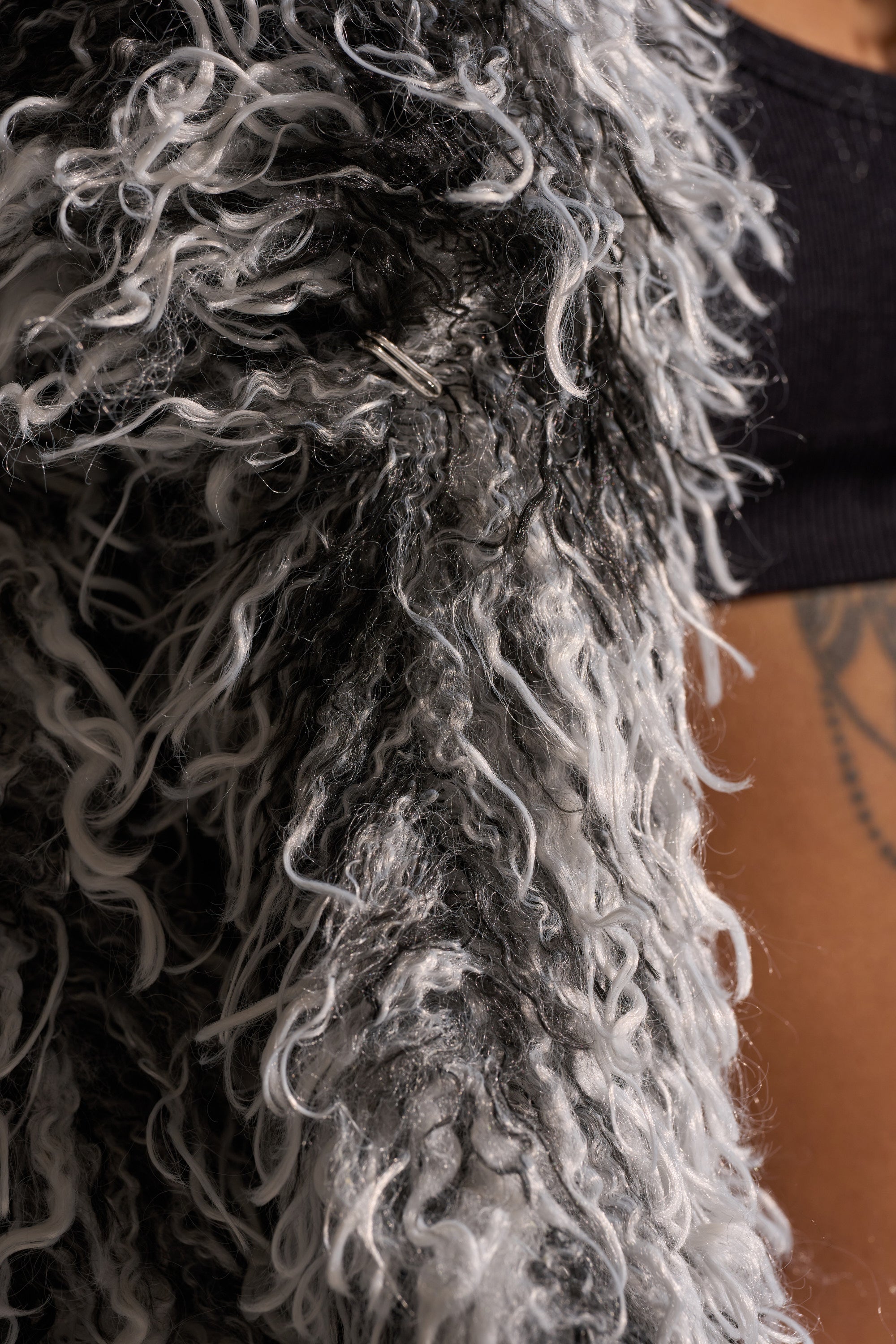 Close-up of the RENDEZVOUS SHAGGY FAUX FUR coat featuring curly, poodle-like texture, worn over a black crop top. A tattooed torso is partially visible in the background.