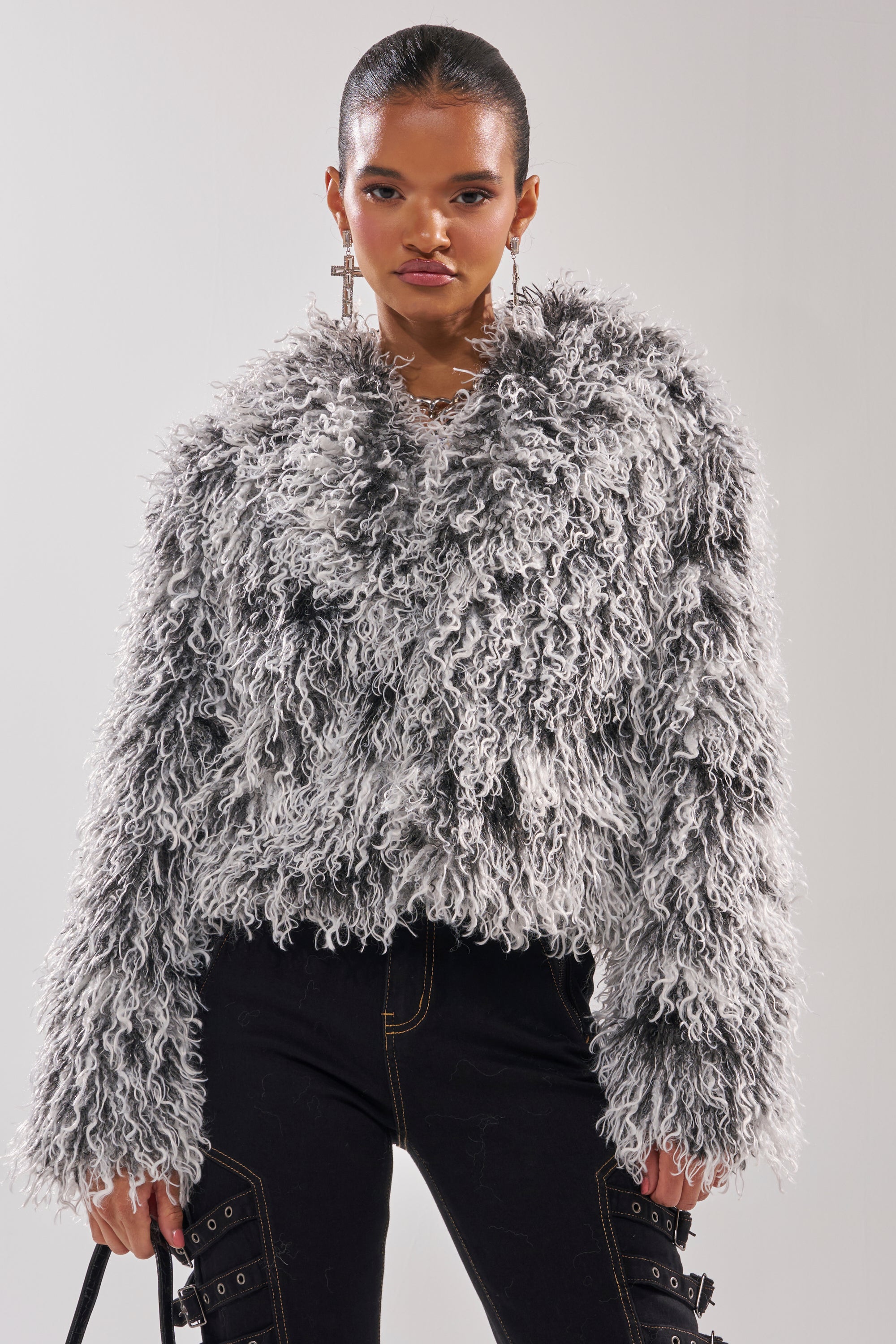 A woman with slicked-back hair models the RENDEZVOUS SHAGGY FAUX FUR cropped coat in gray and black, paired with black pants featuring visible stitching and buckles, plus large cross earrings, standing before a plain background. 0