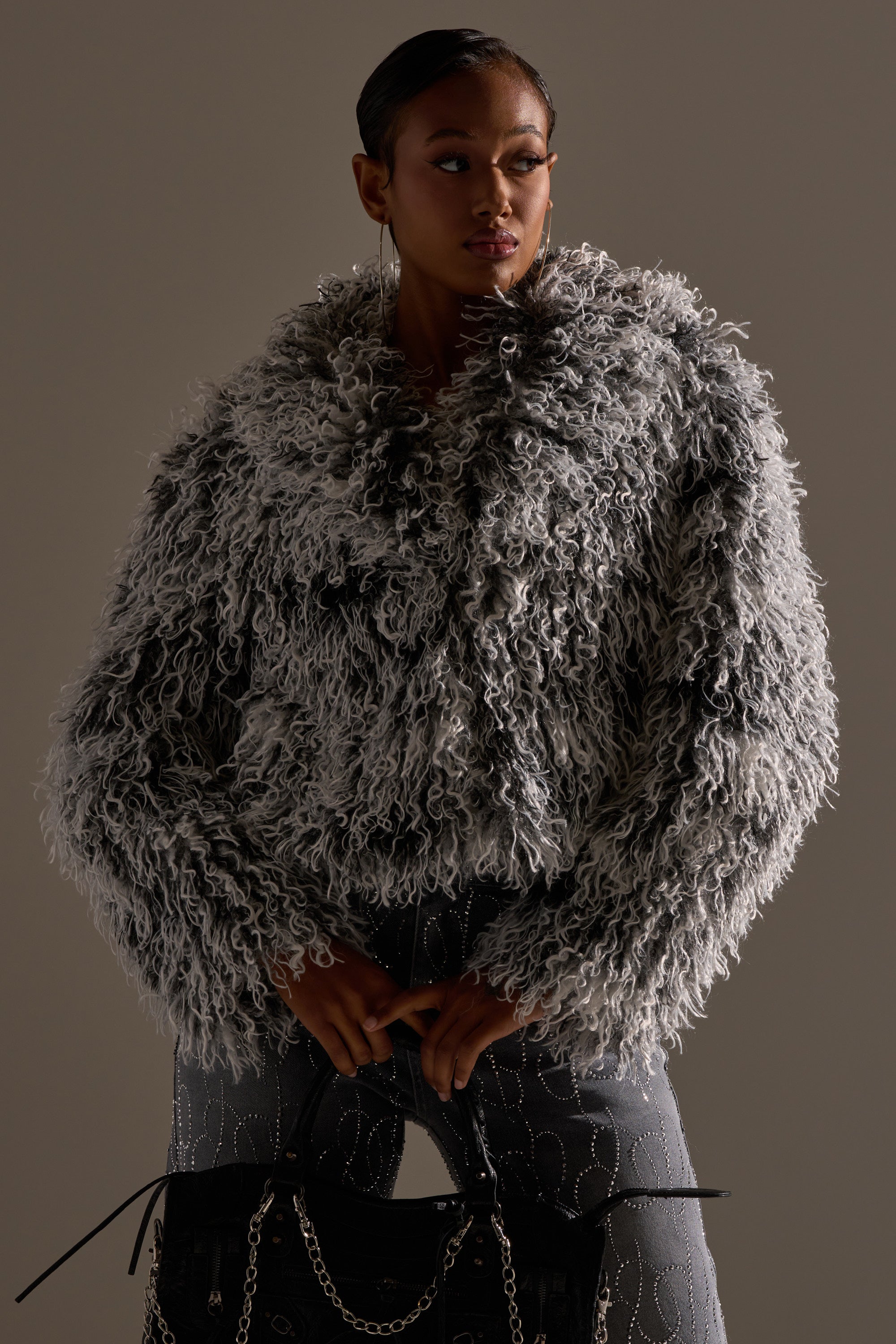 A woman stands confidently in the RENDEZVOUS SHAGGY FAUX FUR coat, holding a black chain-detail handbag and looking poised to the side against a neutral background.