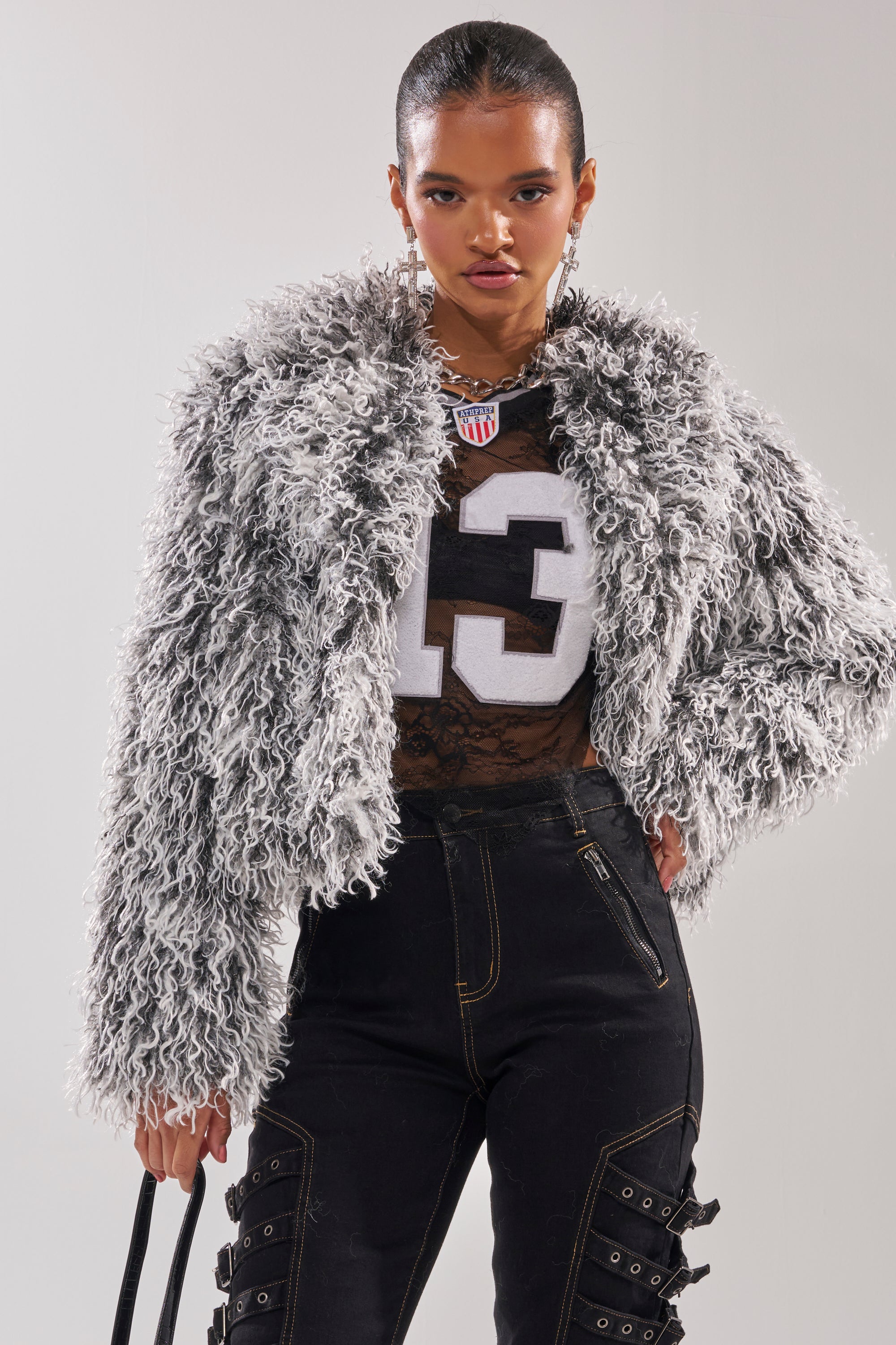 A woman with slicked-back hair wears the RENDEZVOUS SHAGGY FAUX FUR coat over a sheer black top with "13", paired with high-waisted dark pants featuring zippers and straps, and carries a chic black handbag. 1