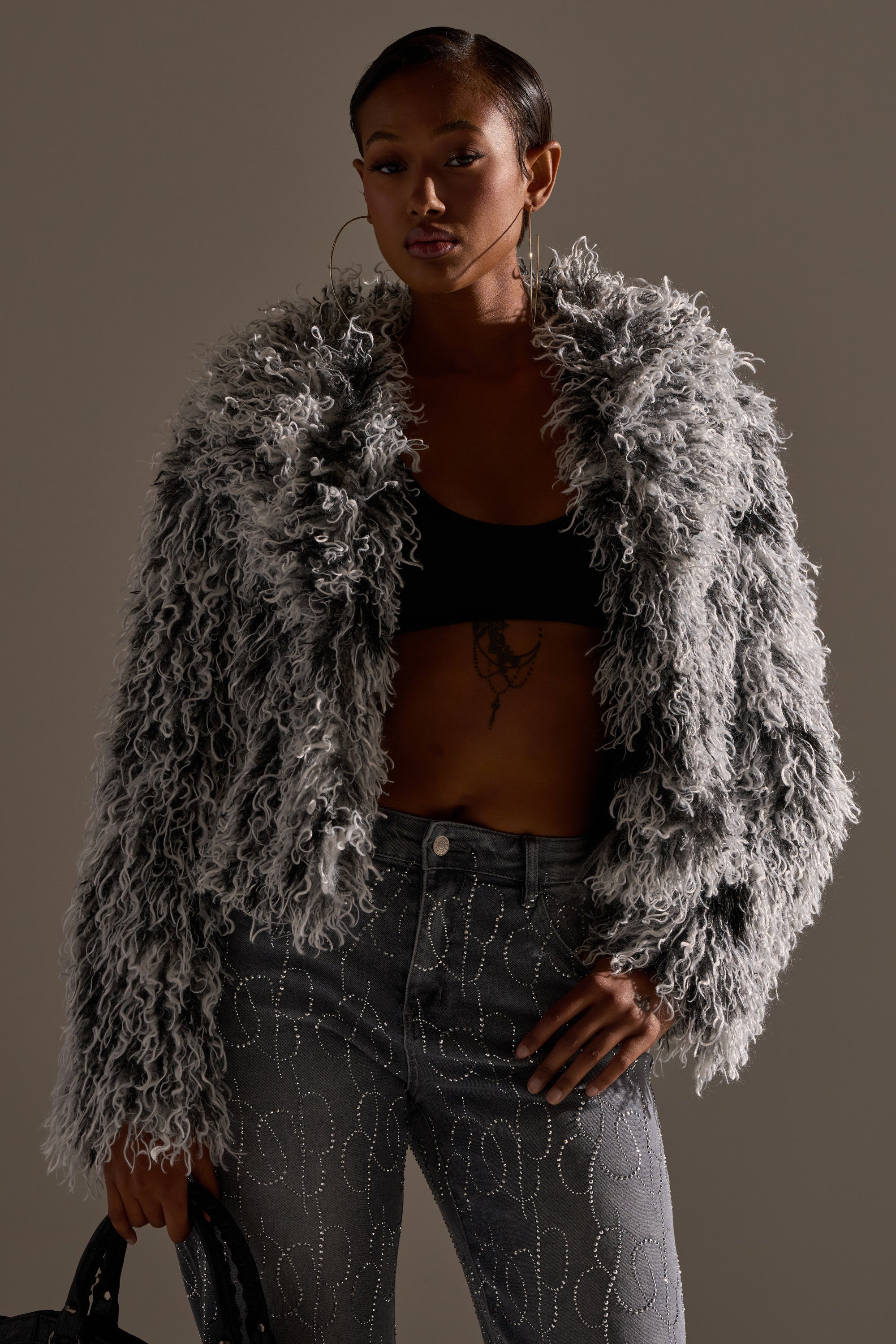 A person poses confidently in the RENDEZVOUS SHAGGY FAUX FUR coat over a black crop top and high-waisted gray embroidered jeans, holding a black handbag against a plain background.