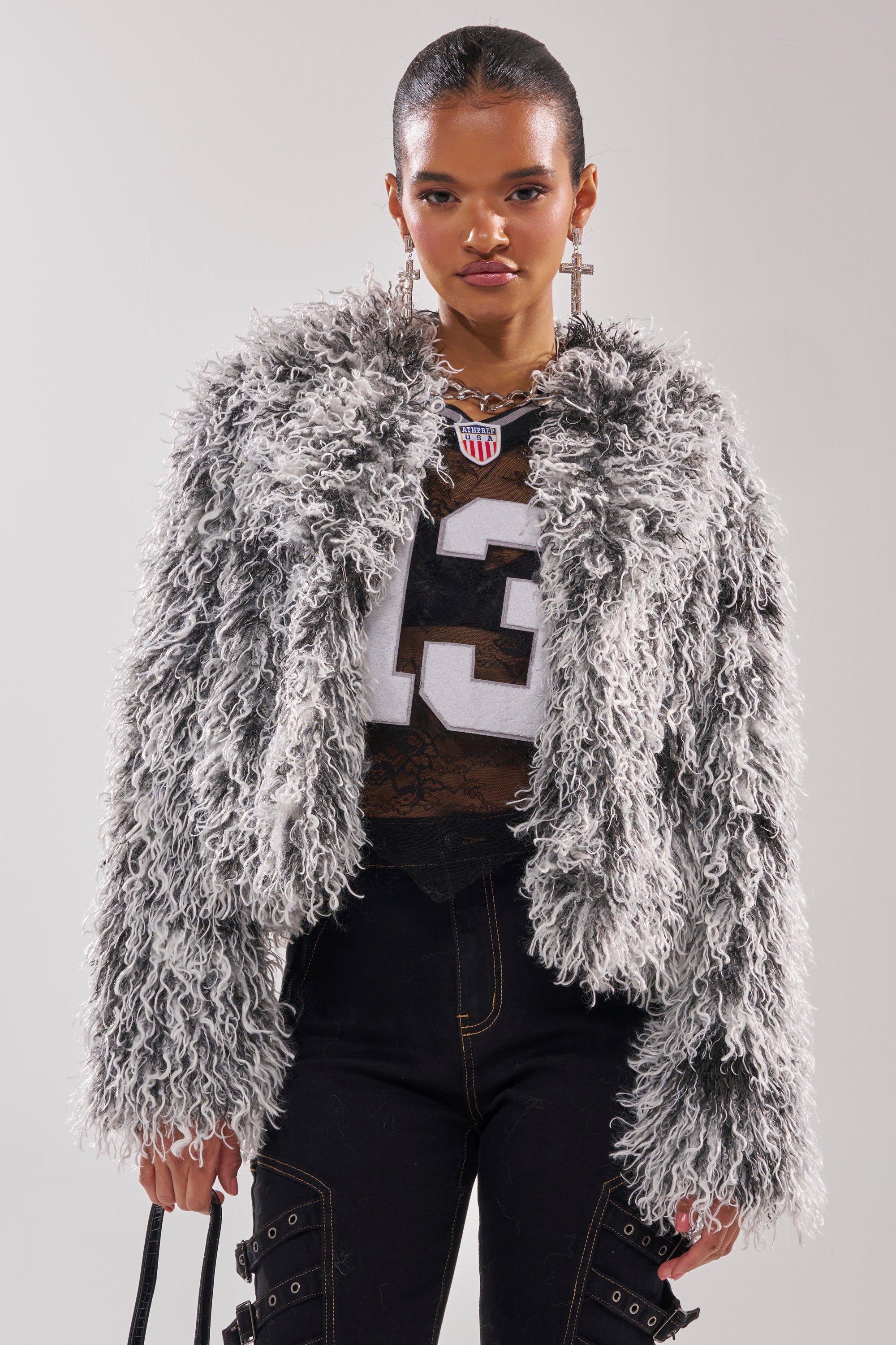 Wearing the RENDEZVOUS SHAGGY FAUX FUR coat over a sheer football jersey with the number 13, black strap pants, statement earrings, and carrying a black bag, a person poses against a plain background. 2
