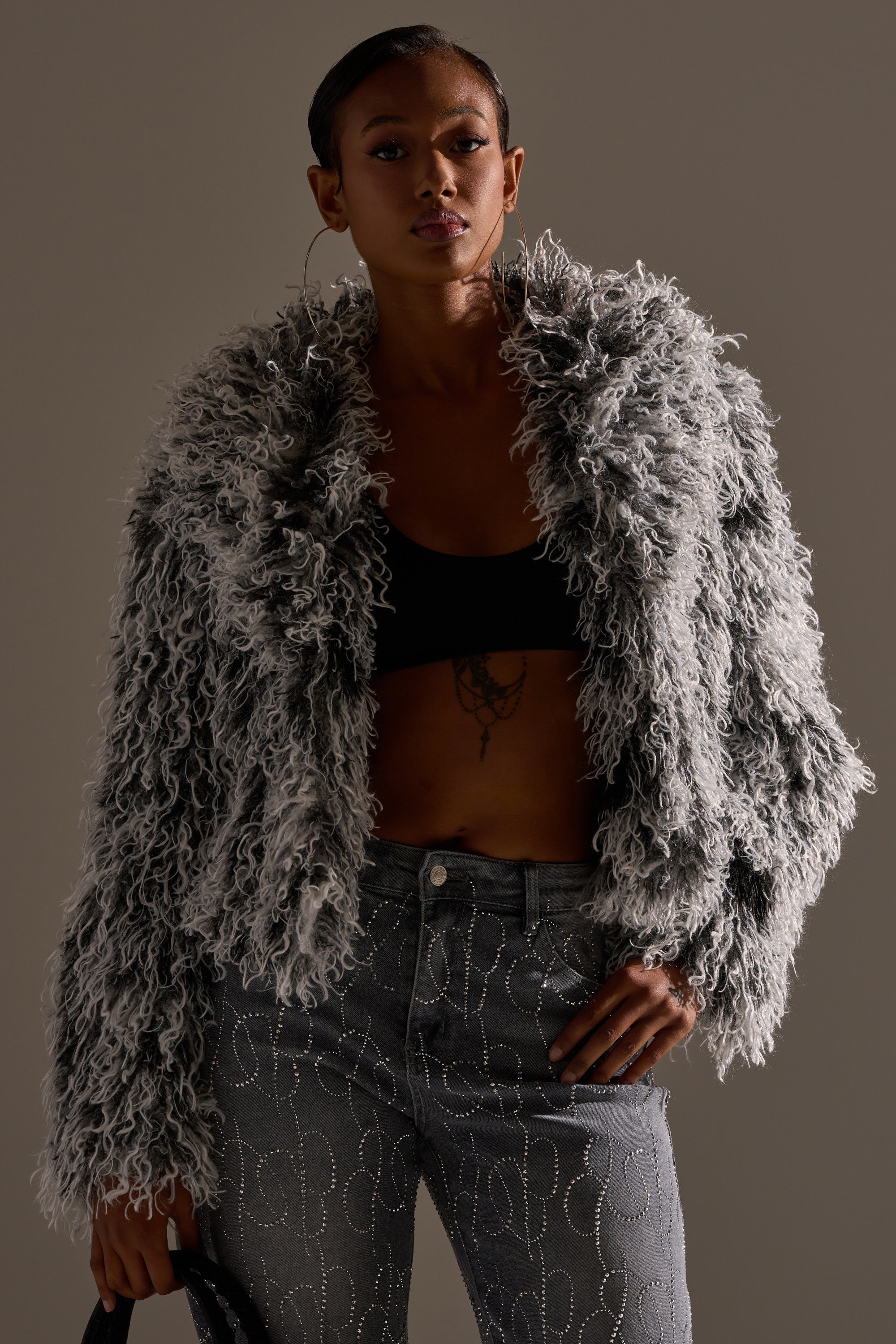 A person wears the RENDEZVOUS SHAGGY FAUX FUR jacket, hoop earrings, a black crop top, and high-waisted patterned jeans, standing confidently against a plain background.