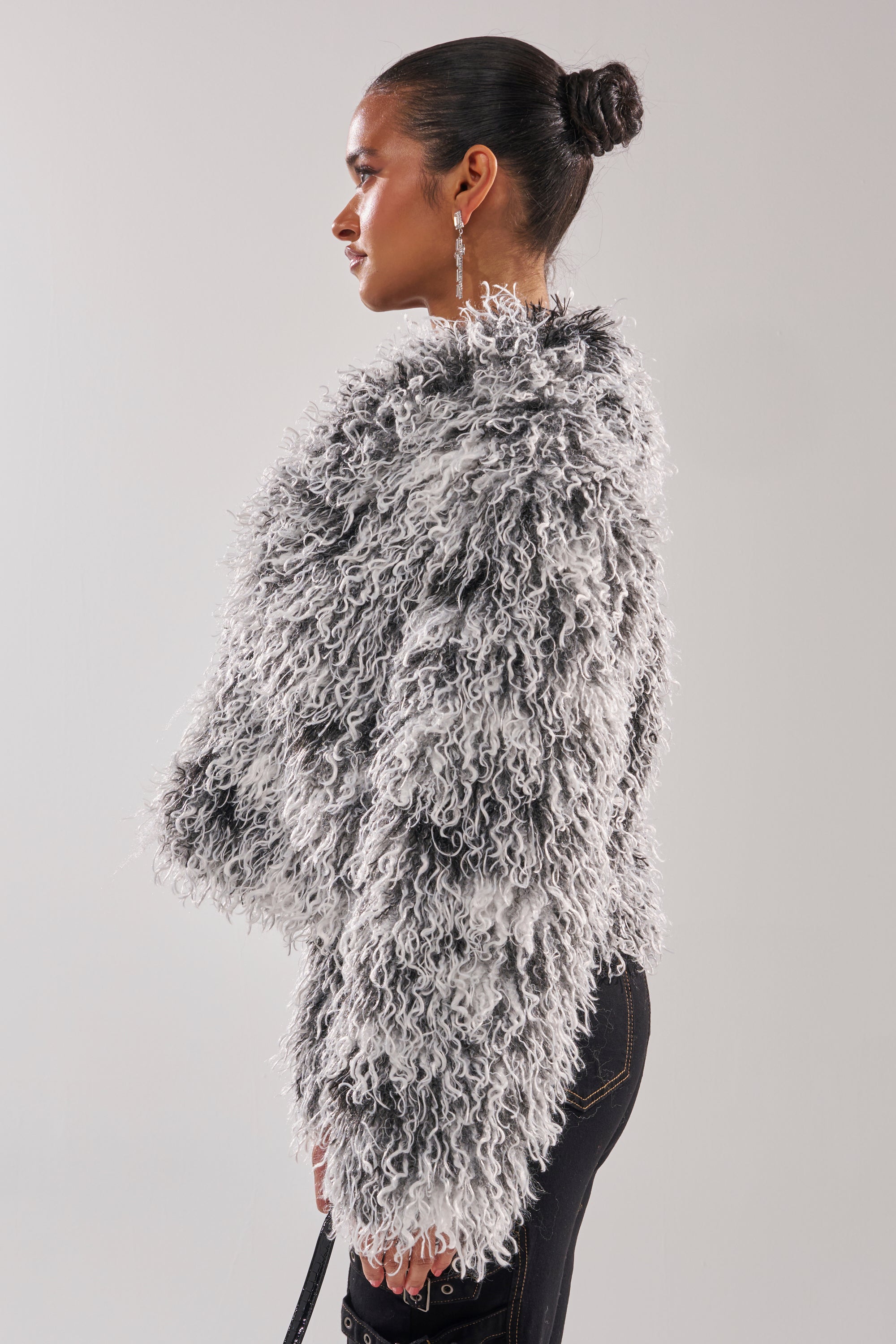 A woman with her hair in a bun wears the RENDEZVOUS SHAGGY FAUX FUR black-and-white textured jacket and dark jeans, standing in profile against a plain, light background. 3