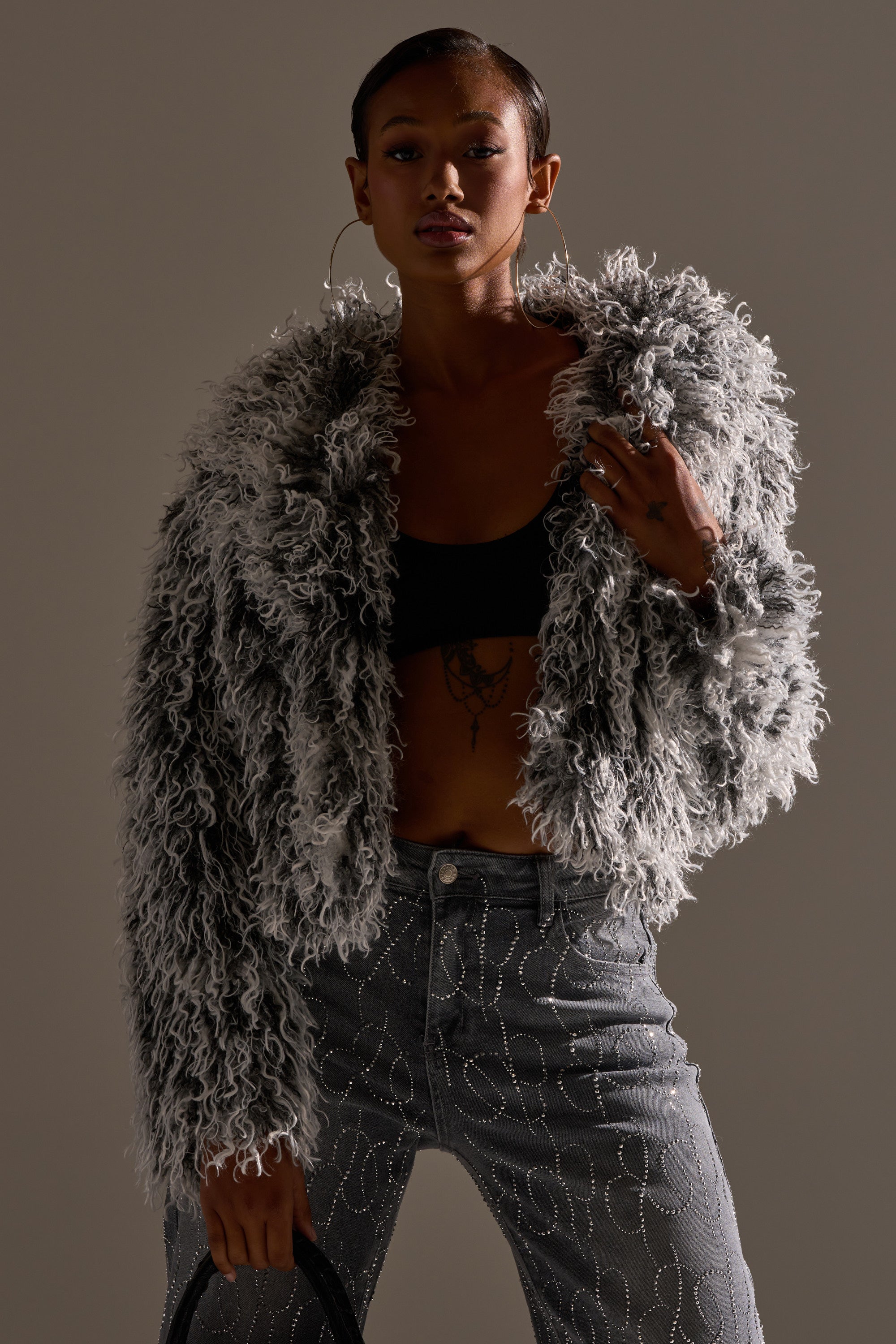 A person poses confidently in the RENDEZVOUS SHAGGY FAUX FUR jacket over a black crop top, paired with hoop earrings and high-waisted gray jeans featuring swirling white patterns.