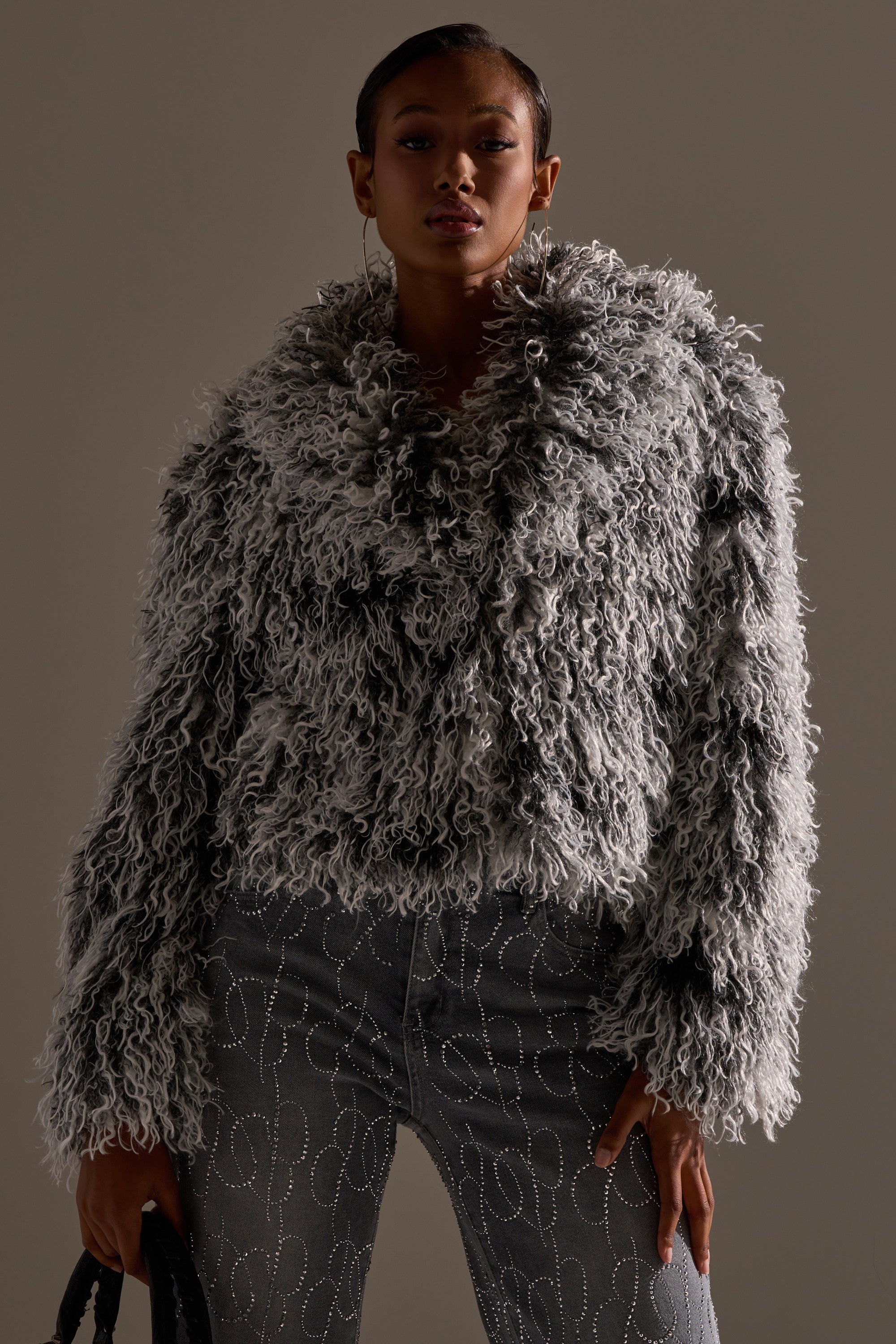Wearing the RENDEZVOUS SHAGGY FAUX FUR cropped coat over embellished jeans, a person stands against a plain background, holding a black bag at their side.