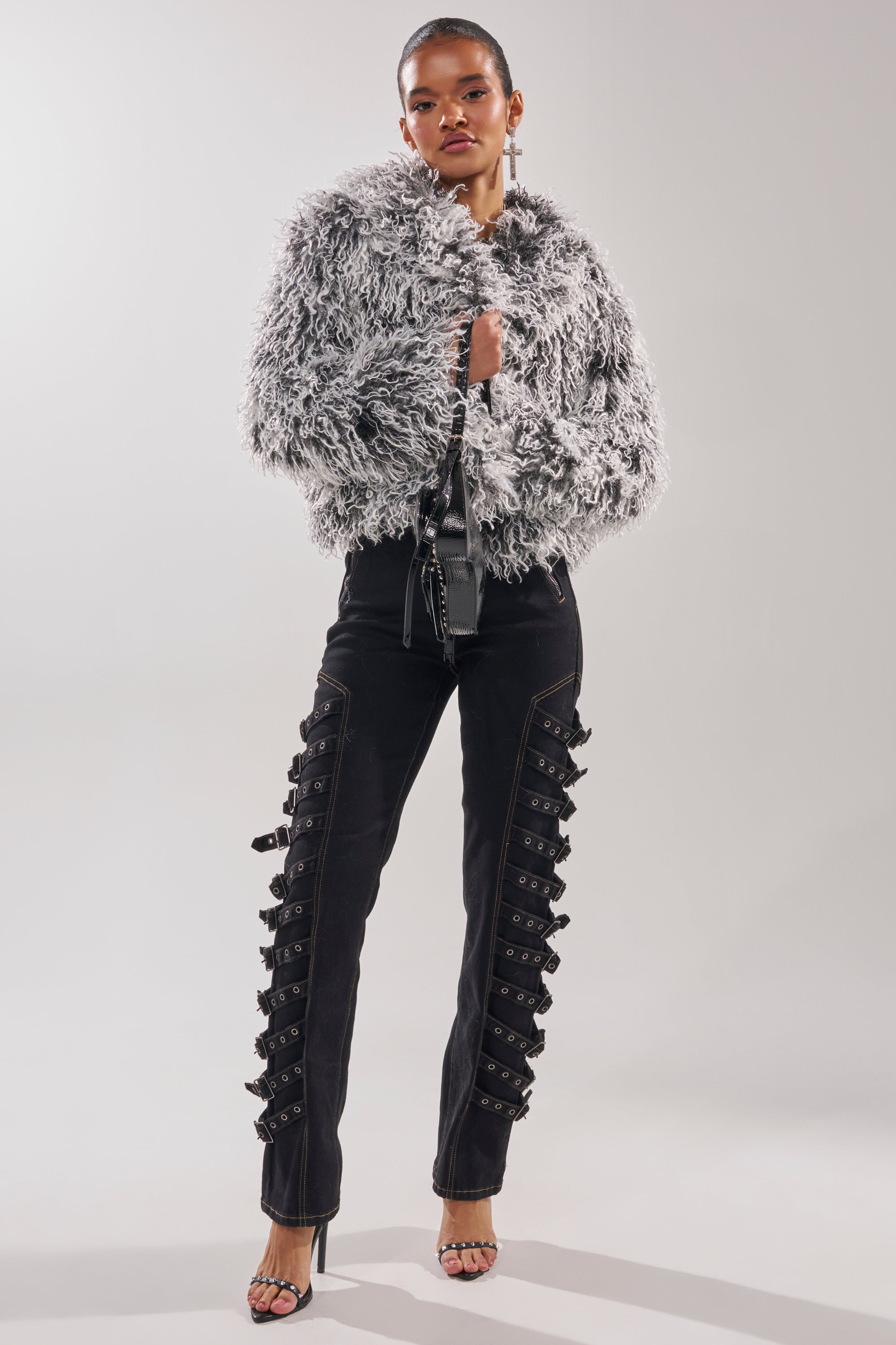 Wearing the RENDEZVOUS SHAGGY FAUX FUR coat, black pants with side buckles, black sandals, and long earrings, a person poses confidently against a plain background. 5