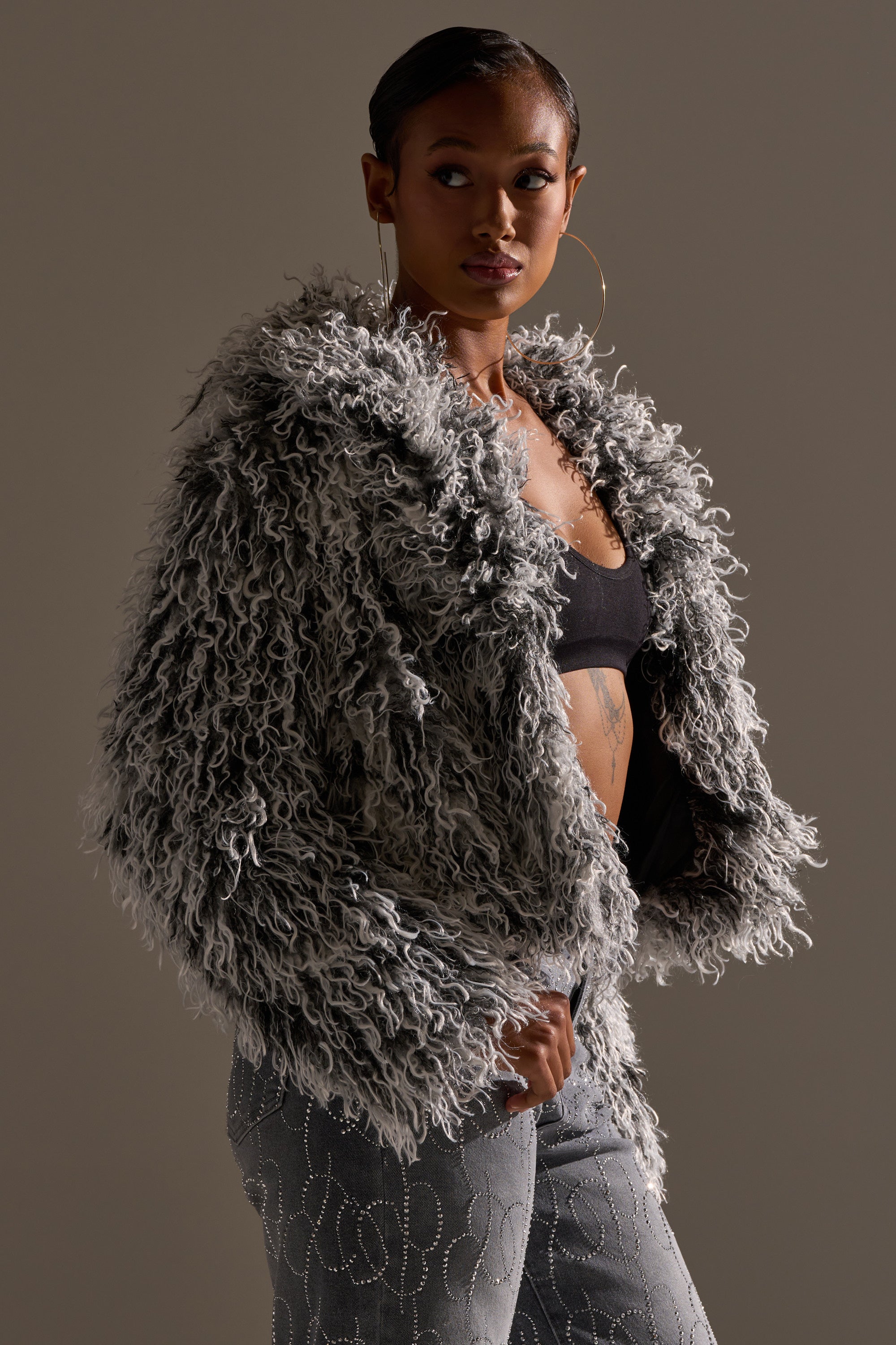 A woman models the RENDEZVOUS SHAGGY FAUX FUR coat over a black top with hoop earrings and patterned pants, set against a neutral background.