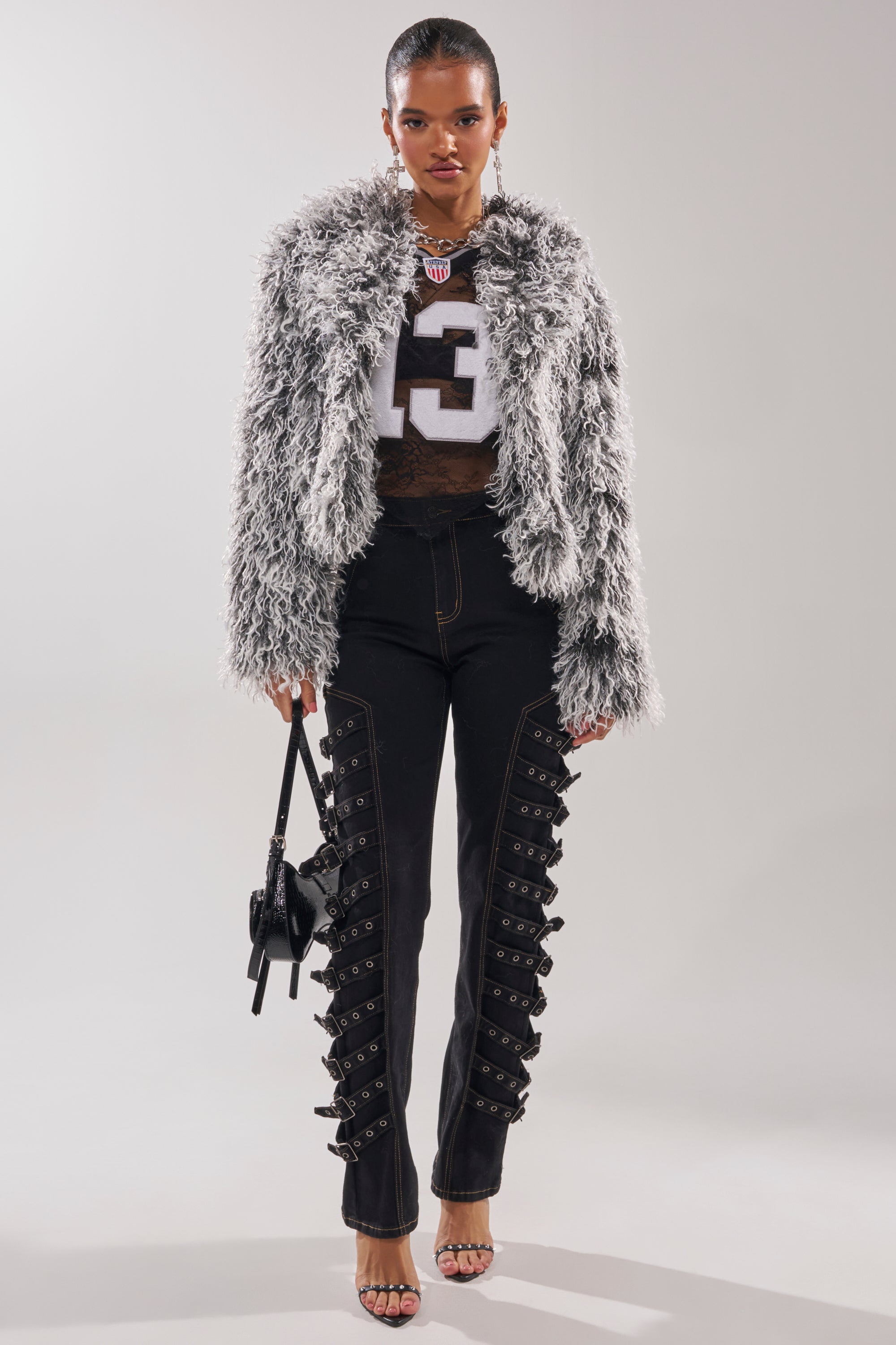 A woman wearing the RENDEZVOUS SHAGGY FAUX FUR coat with an oversized collar, a cropped sports jersey, black buckle-detail pants, strappy heels, and a small handbag poses against a plain light background. 6