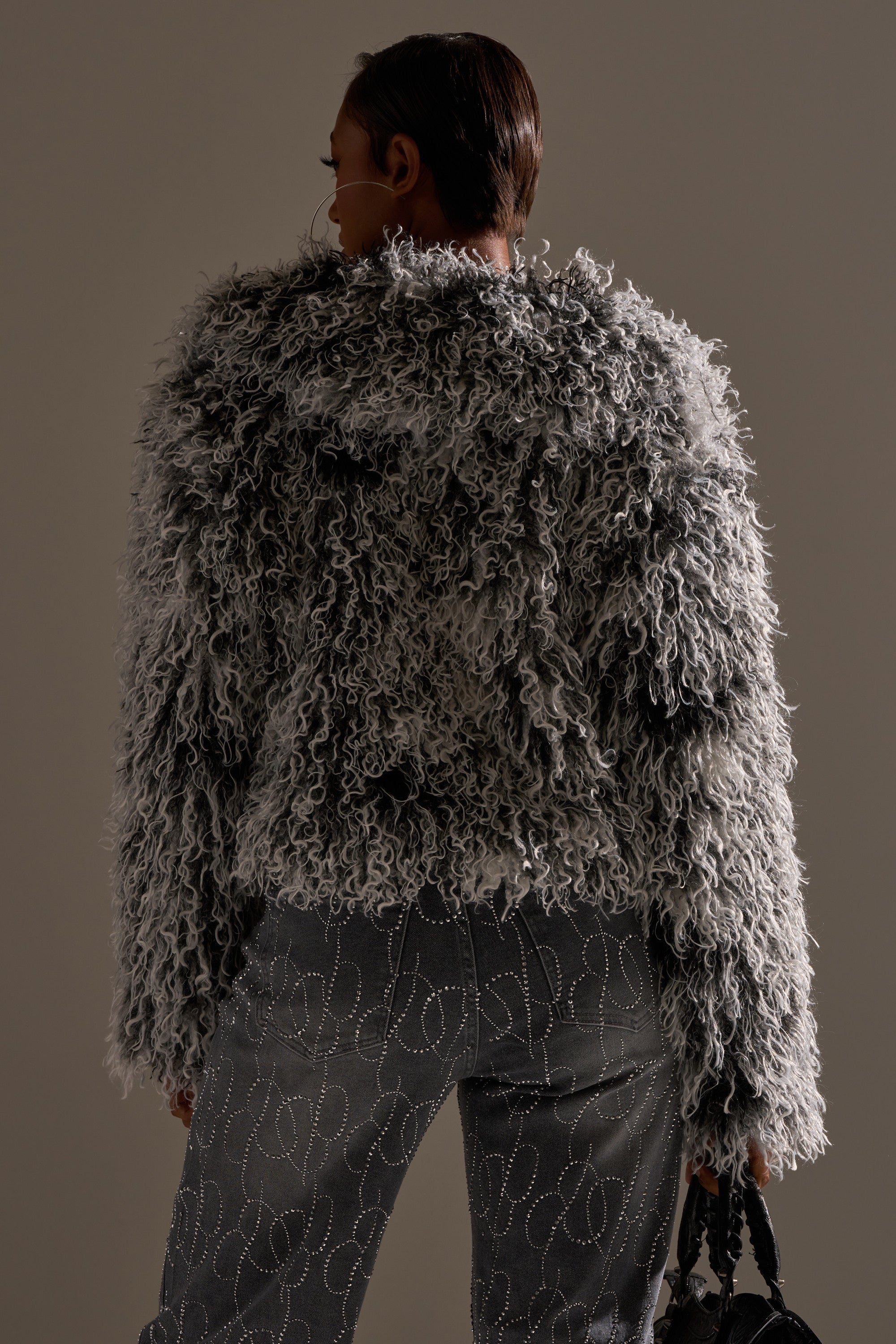 A person stands facing away, wearing the RENDEZVOUS SHAGGY FAUX FUR jacket in gray and embellished gray jeans. Soft lighting emphasizes the jacket’s curly texture as they hold a small black bag in their left hand.