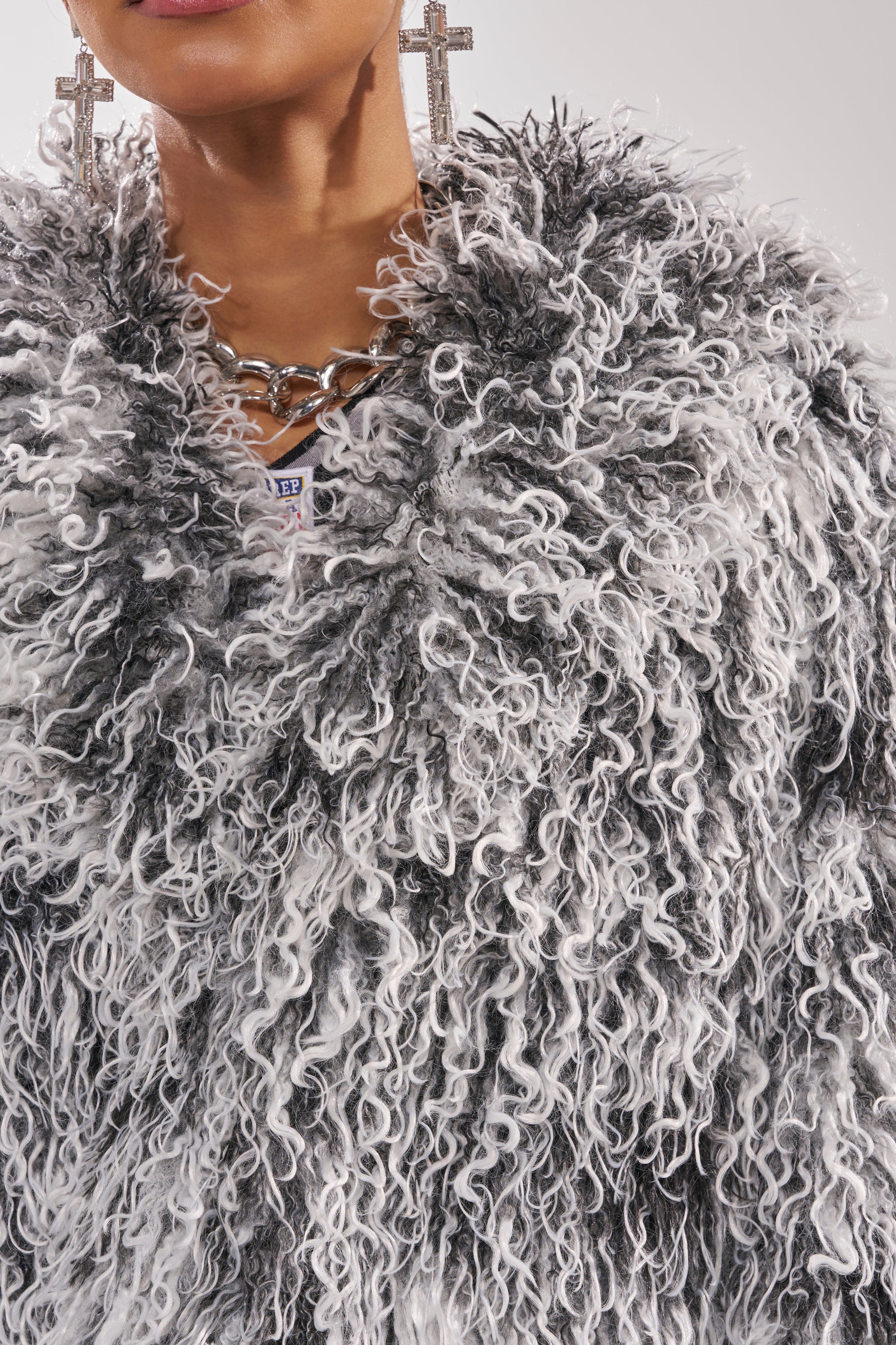 Wearing the RENDEZVOUS SHAGGY FAUX FUR coat with an oversized collar, plus a chunky chain necklace and large cross-shaped earrings, only the lower face and upper torso are shown. 7