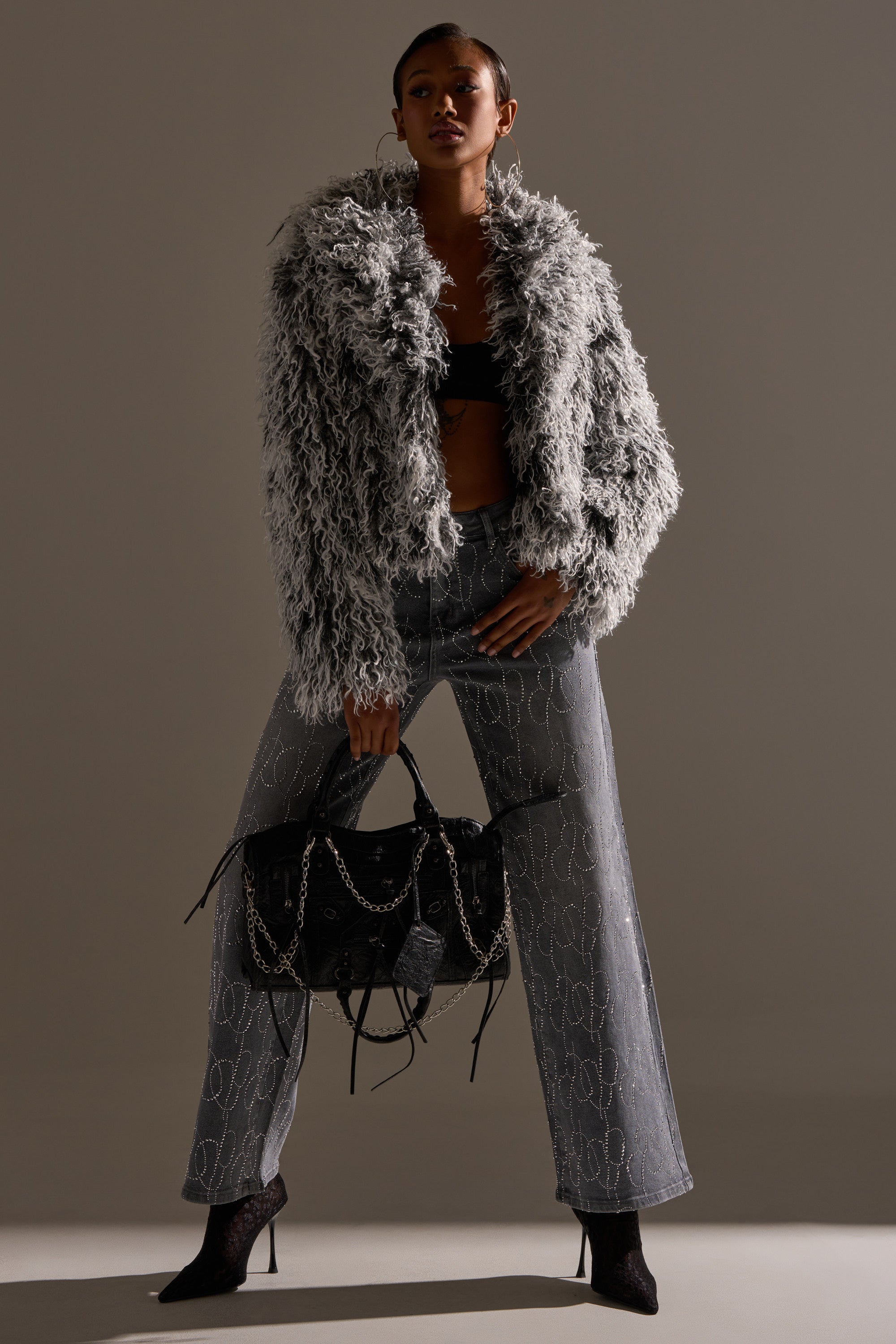 A person poses confidently in the RENDEZVOUS SHAGGY FAUX FUR coat, paired with a black top, gray wide-leg patterned pants, black heeled boots, and a large black handbag against a softly lit background.
