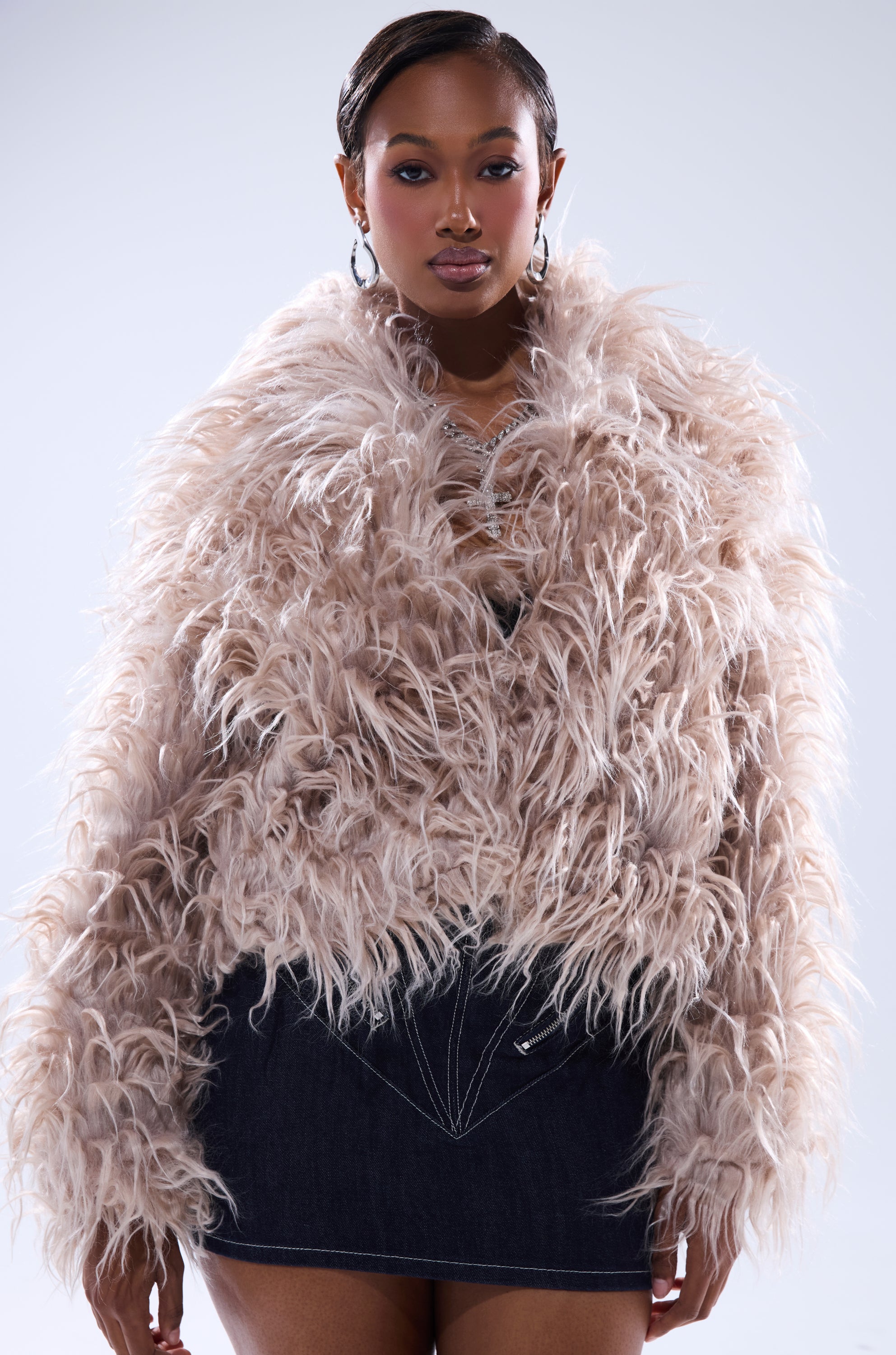 A woman exudes confidence in the MISS ME FAUX FUR, an oversized champagne-colored jacket, styled over a dark denim mini skirt with hoop earrings and sleek side-parted hair against a light background.