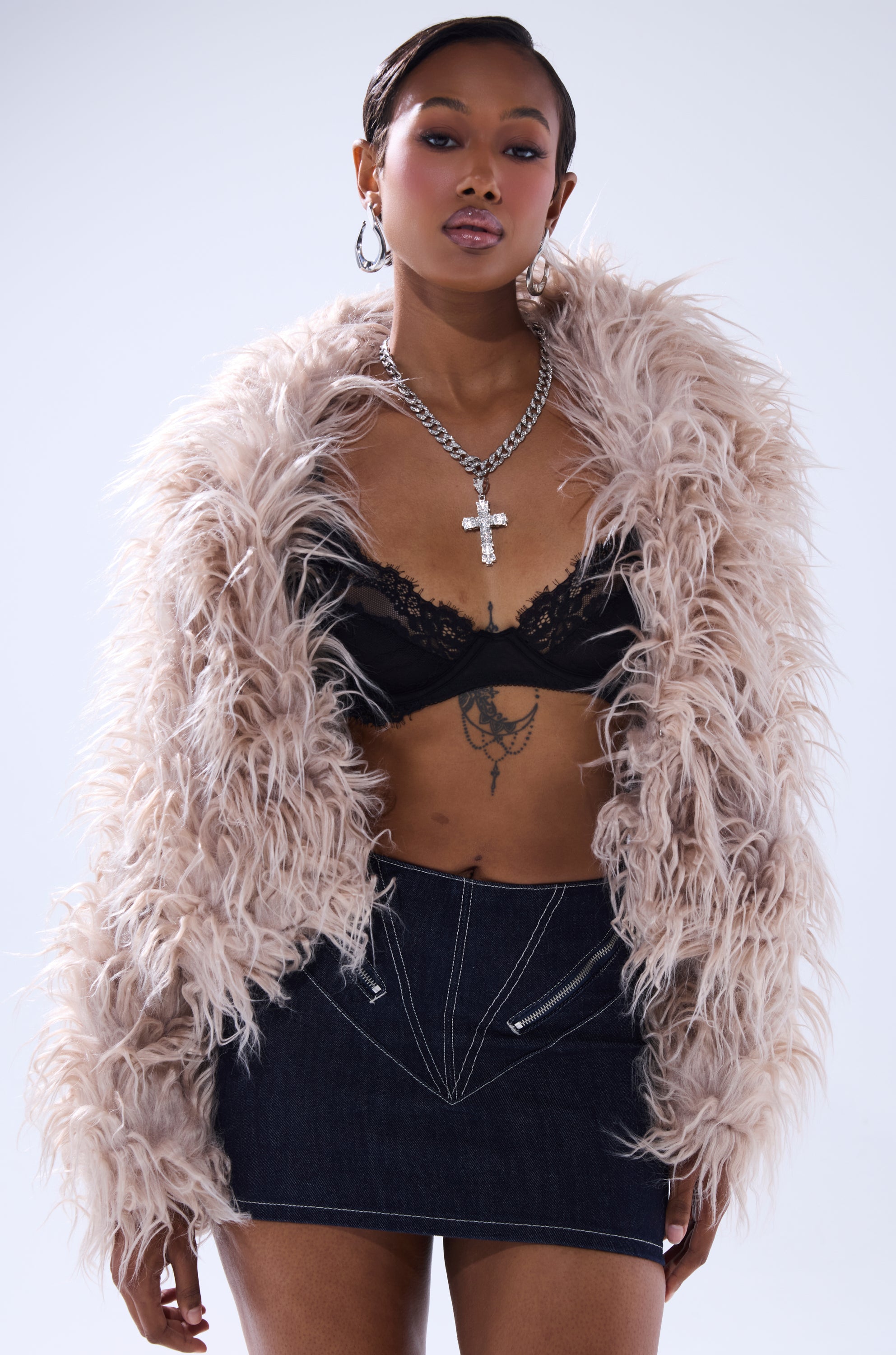 A stylish woman poses in the MISS ME FAUX FUR light pink jacket, black lace bralette, and dark denim mini skirt. She accessorizes with a silver cross necklace, hoop earrings, and shows off a tattoo on her torso.