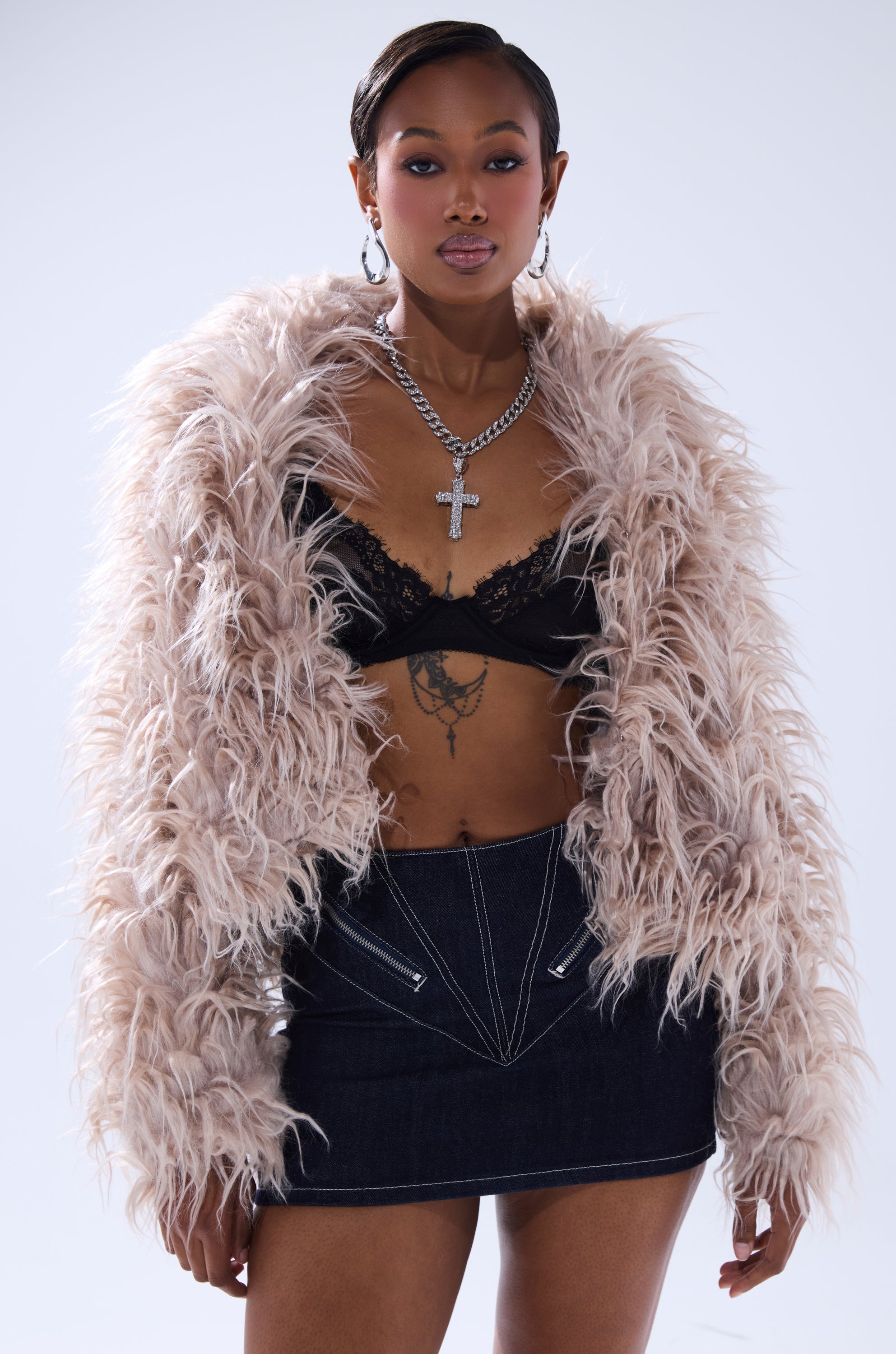 A confident woman poses against a light background, wearing the MISS ME FAUX FUR champagne jacket over a black lace bralette, paired with a dark denim mini skirt, hoop earrings, layered necklaces, and showcasing a torso tattoo.