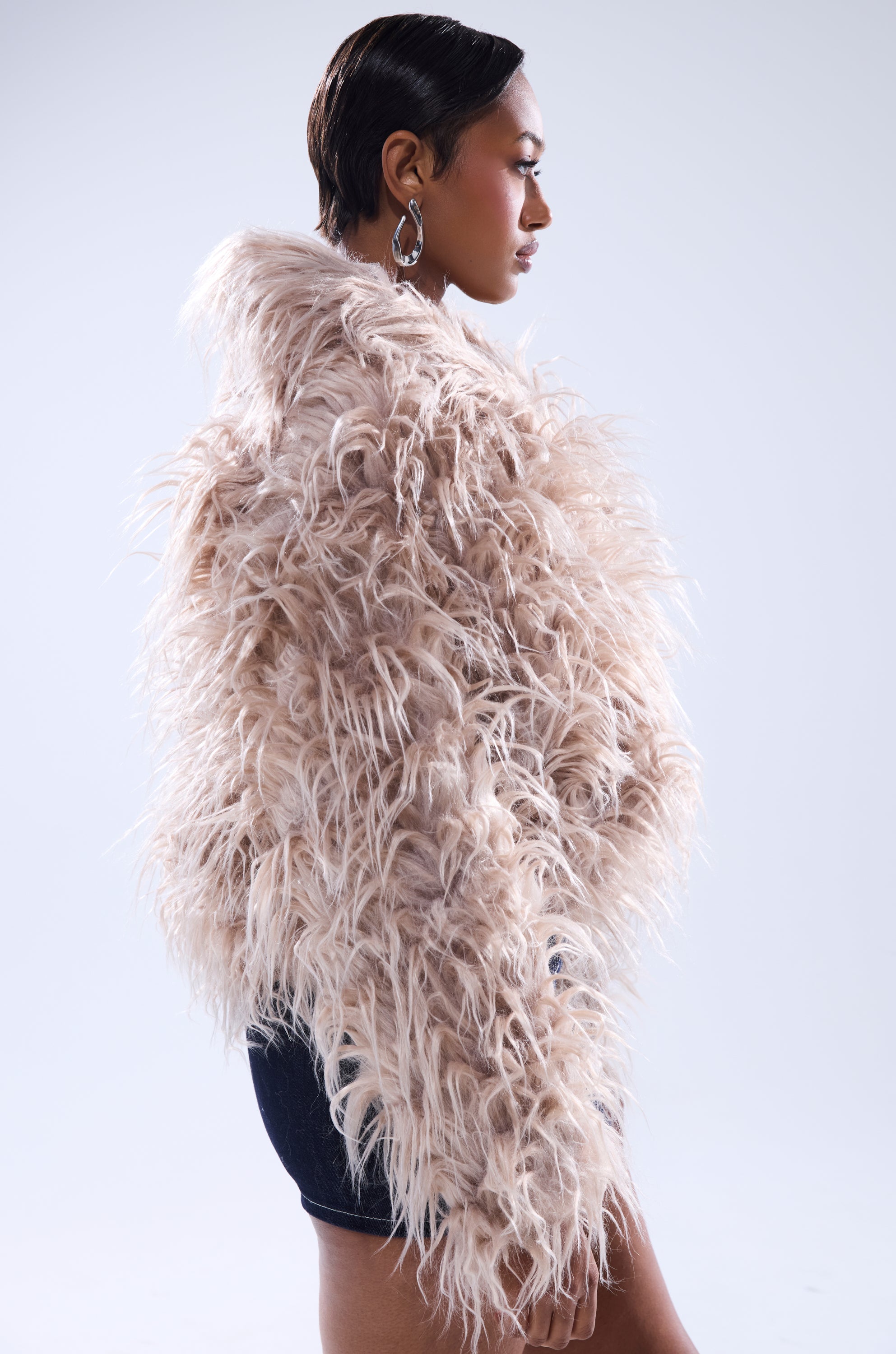 A woman in profile models the MISS ME FAUX FUR jacket—an oversized, light faux fur piece—paired with large hoop earrings and dark shorts, her short sleek hair standing out against a plain white backdrop.