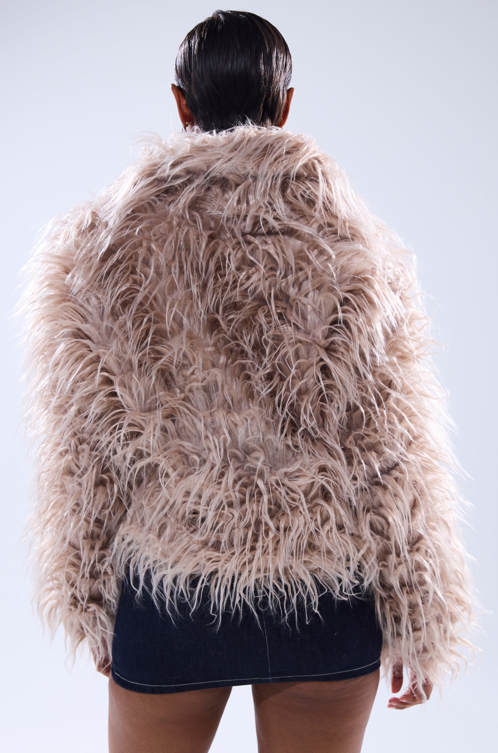 A person with short, slicked-back hair is seen from behind in the oversized beige MISS ME FAUX FUR jacket and a dark denim mini skirt, set against a light background.
