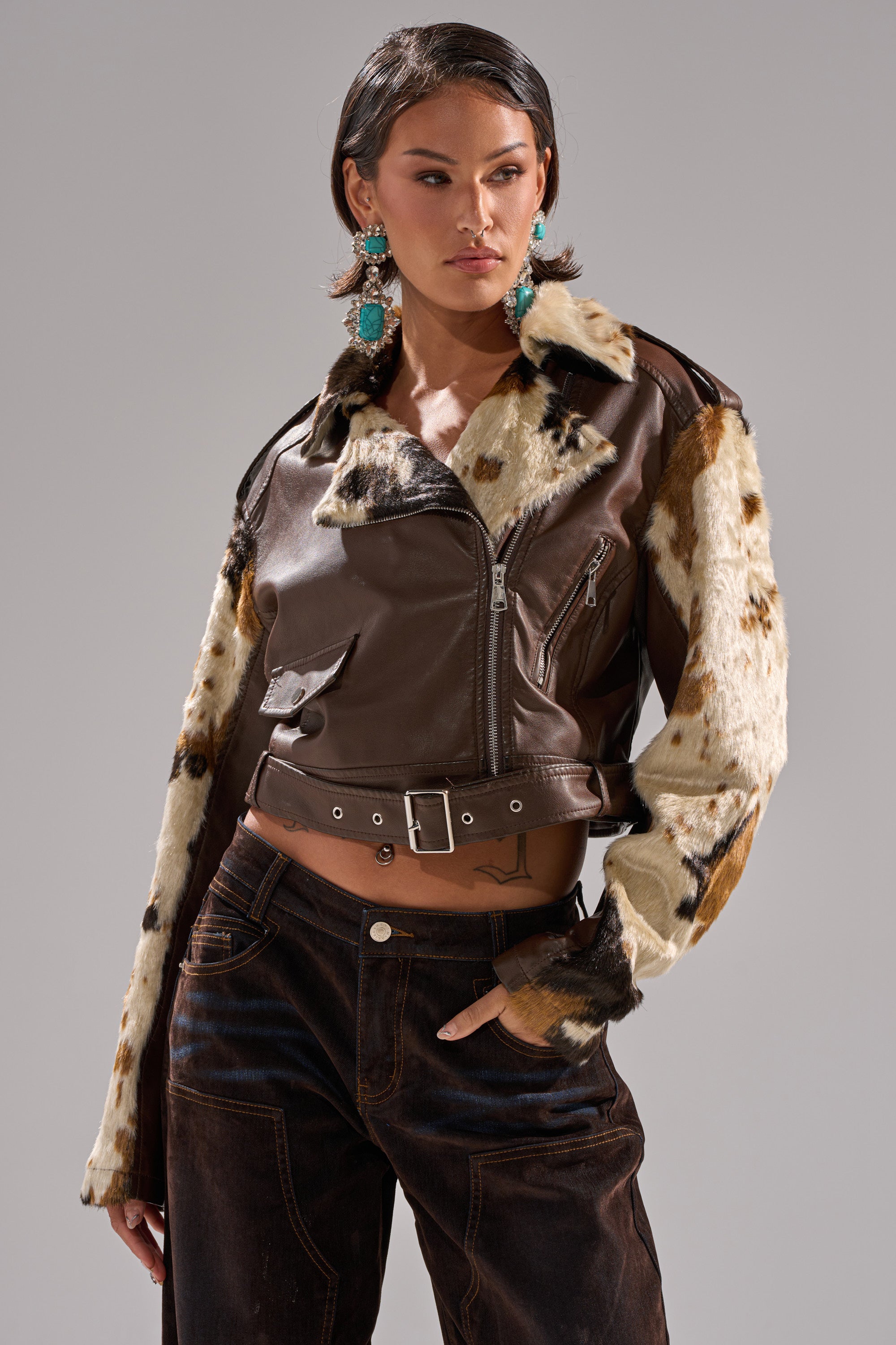 A woman poses in the OUT WEST CROPPED MOTO, a cropped moto jacket featuring a cowhide print fur collar and sleeves. She pairs it with dark pants, large turquoise earrings, and stands against a plain gray background. 0