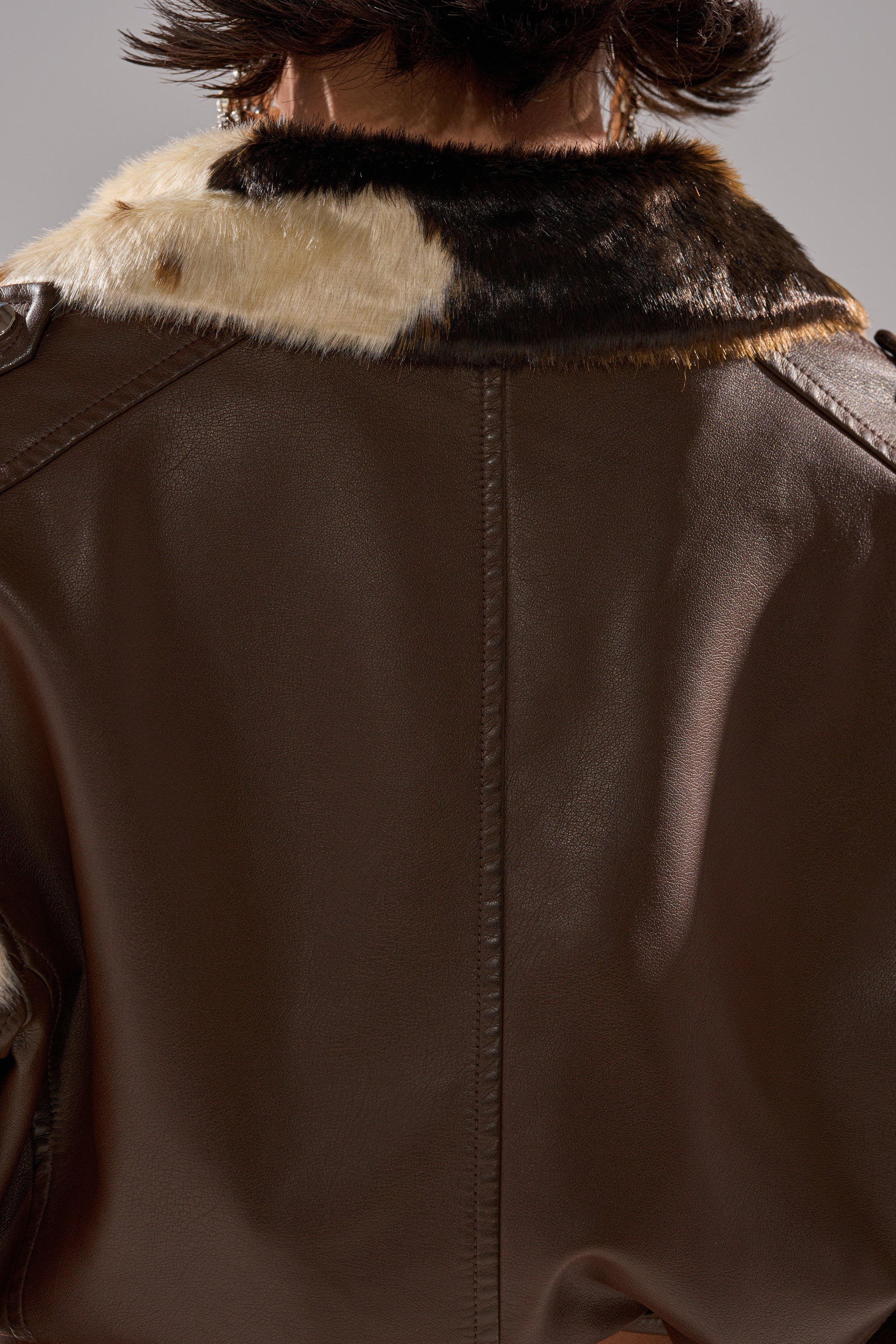 Close-up of the back of a person wearing the OUT WEST CROPPED MOTO, a brown faux leather jacket with a white and dark brown cowhide print fuzzy collar. The person’s short, dark hair is partially visible above the collar. 14