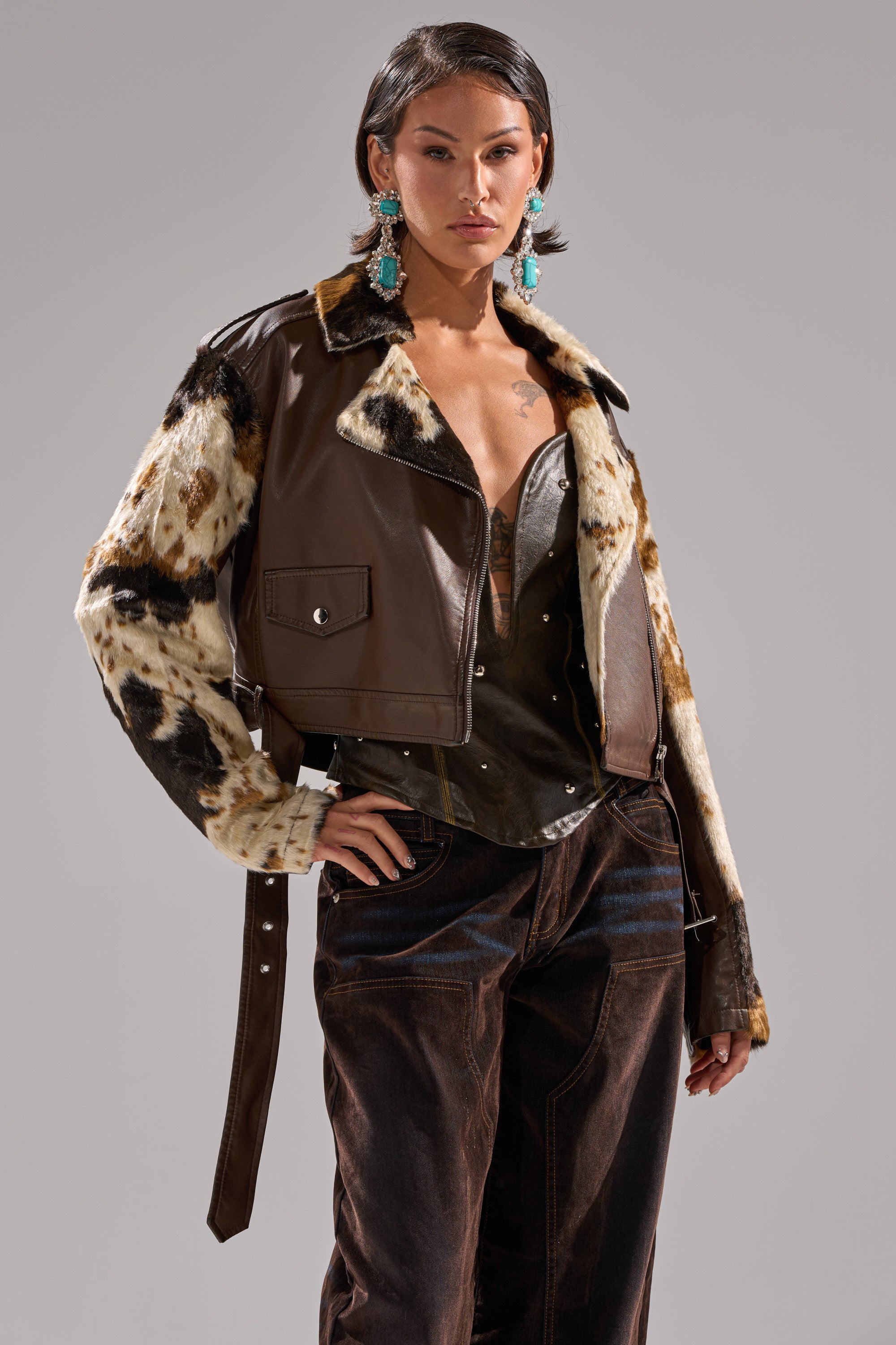 A person with short hair models the OUT WEST CROPPED MOTO cowhide print faux leather jacket, large turquoise earrings, strapless metallic top, and dark high-waisted pants against a plain light gray background. 3