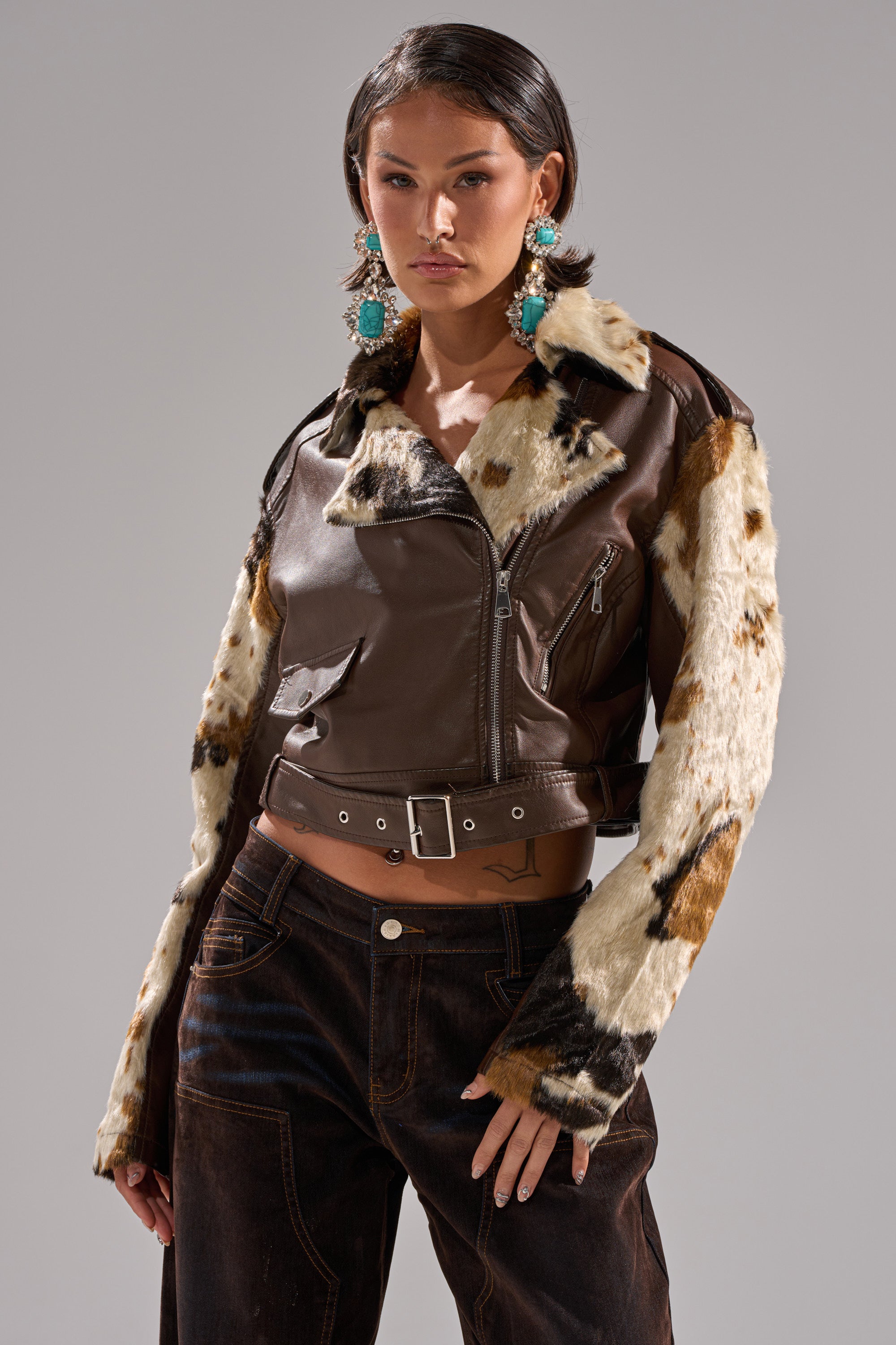 A woman poses confidently in the OUT WEST CROPPED MOTO, a faux leather jacket with cowhide print fur sleeves, paired with dark pants and large turquoise earrings against a neutral background. 5