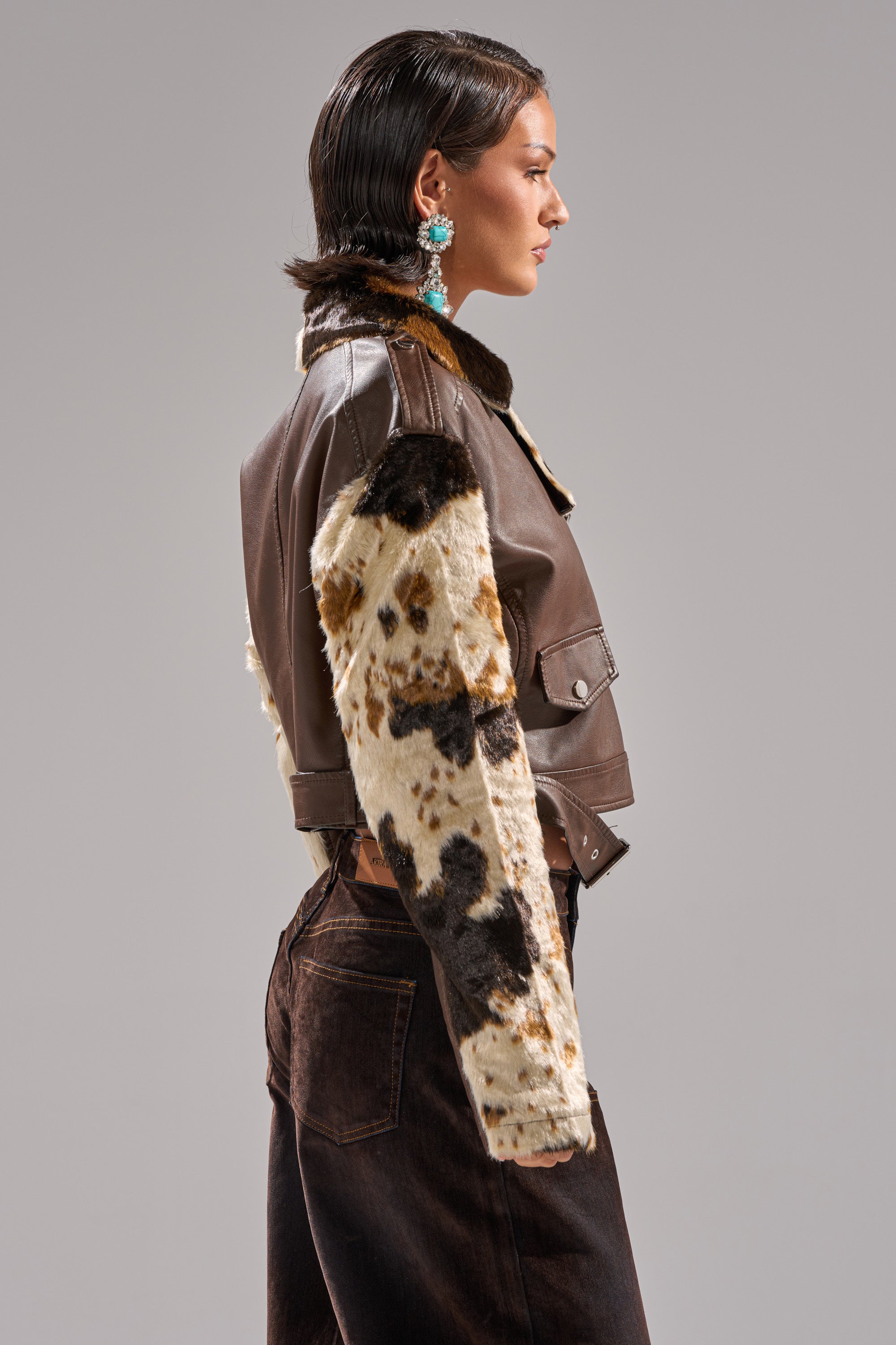A woman in profile wears the OUT WEST CROPPED MOTO, a cropped jacket with fur sleeves in mottled brown and cream cowhide print, paired with dark brown pants and turquoise drop earrings, set against a plain gray background. 6