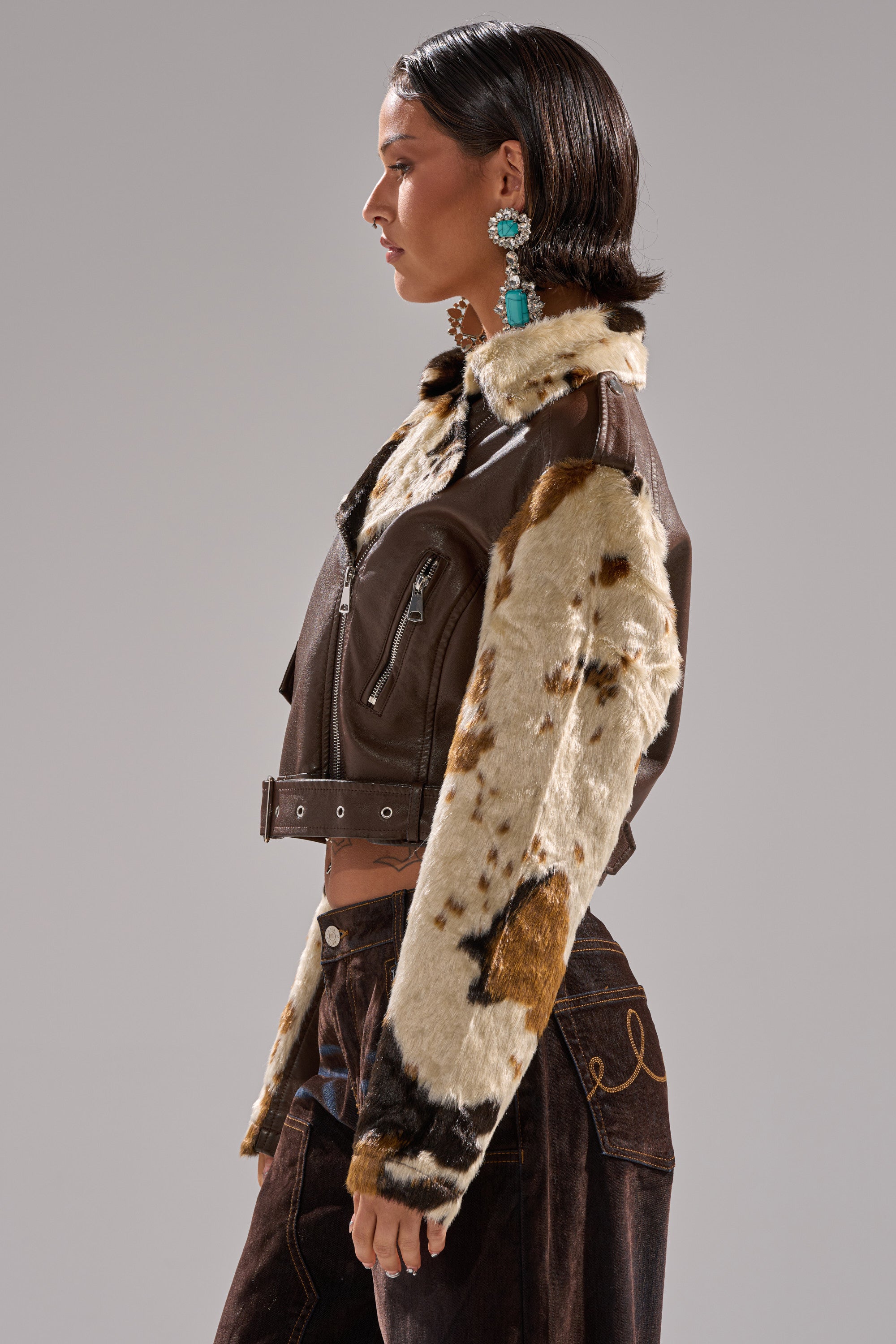 A woman with short dark hair wears the OUT WEST CROPPED MOTO, a cowhide print faux leather jacket with spotted faux fur sleeves, paired with dark brown pants and large statement earrings as she faces left in profile. 7