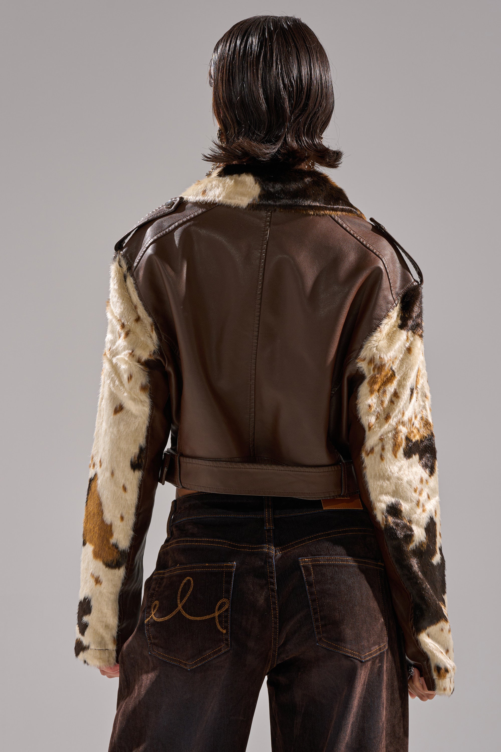 A person with short, dark hair stands facing away, wearing the OUT WEST CROPPED MOTO cowhide print faux leather jacket and dark brown jeans with gold embroidered back pockets. 8