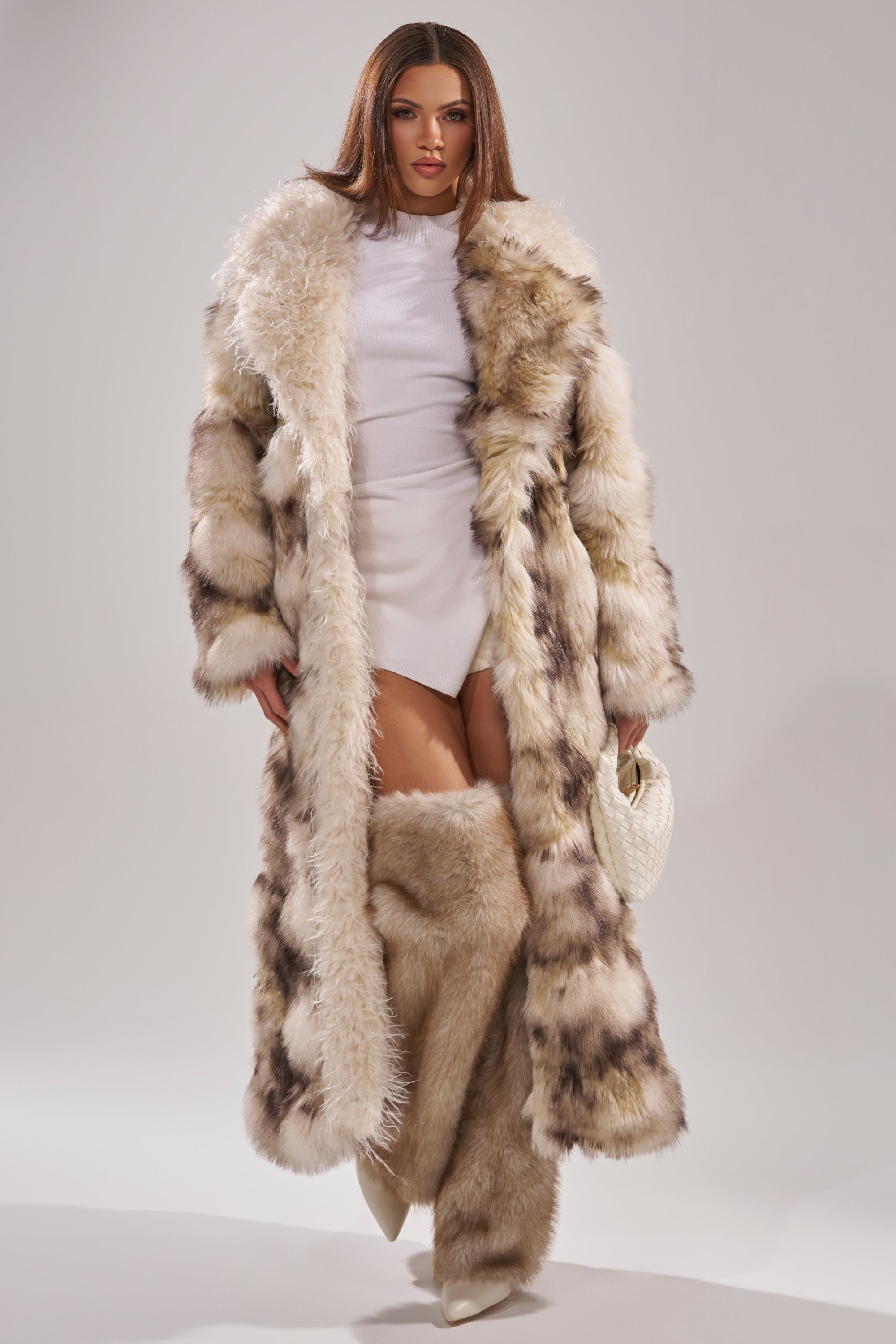 A woman stands confidently in the PARK AVENUE PATCHWORK FAUX FUR coat with matching fur boots, a short white dress, white pointed heels, and a small white handbag, looking directly at the camera against a plain background.