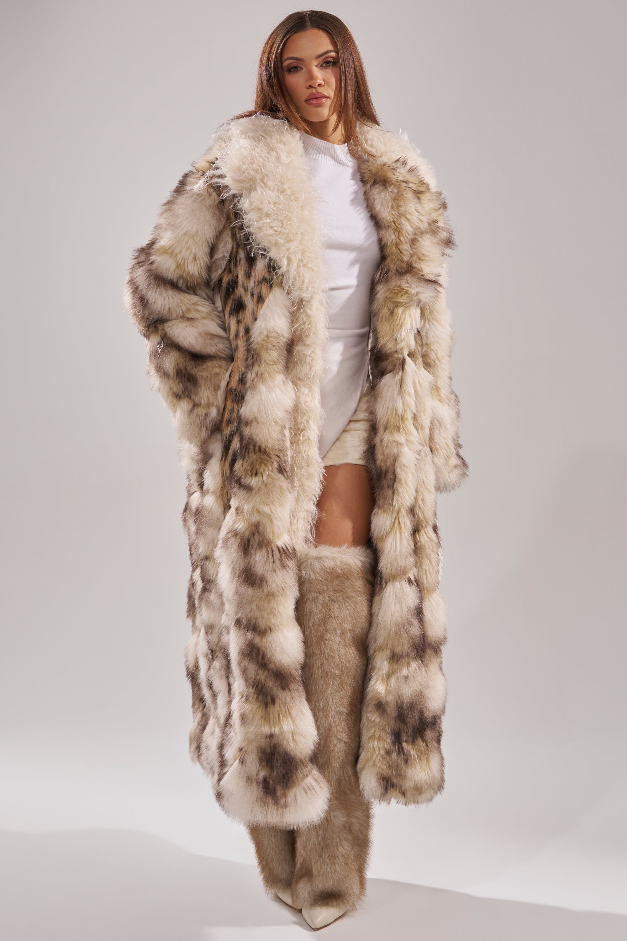 A woman wears the PARK AVENUE PATCHWORK FAUX FUR coat over a white outfit with matching fur boots, posed against a light gray background.