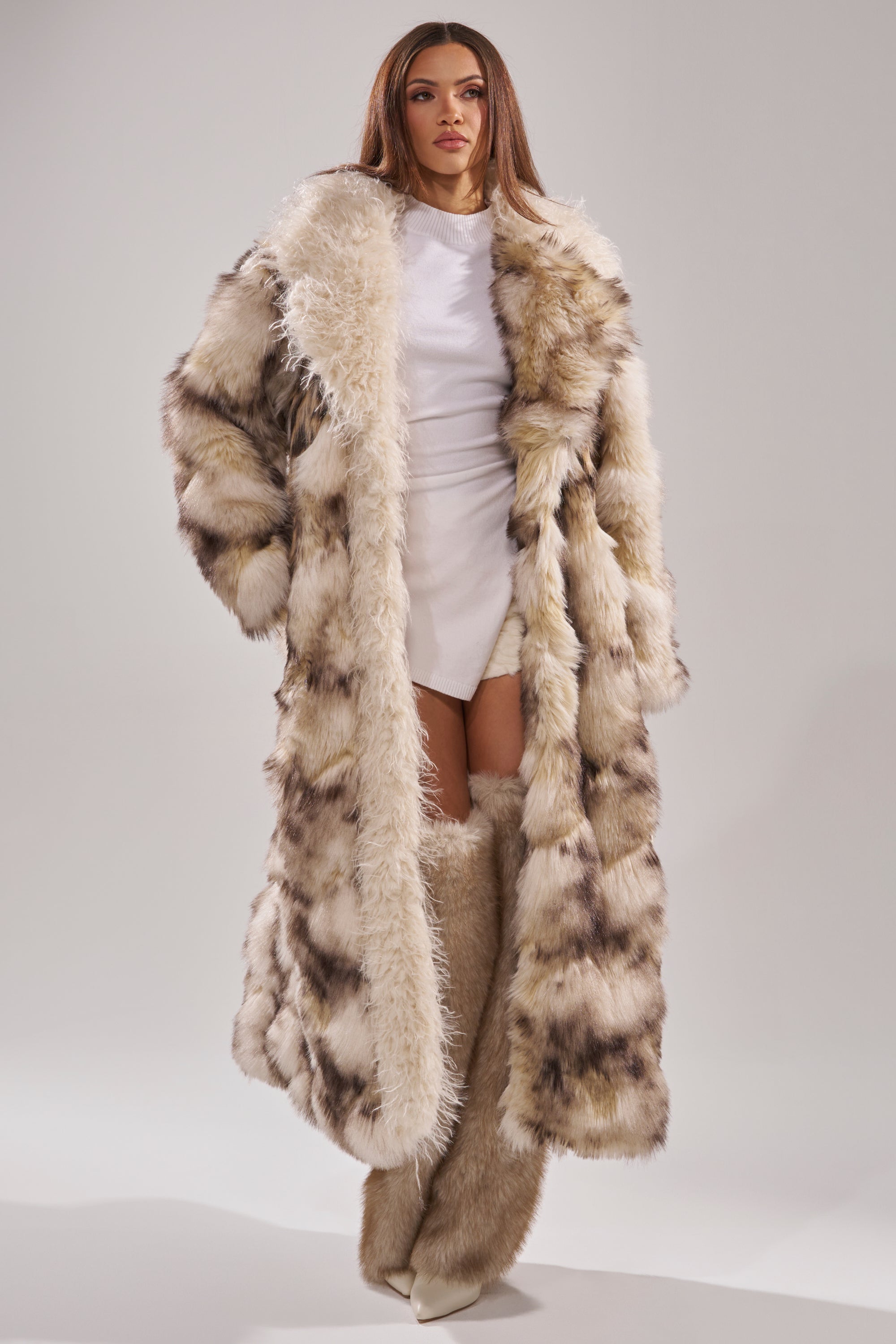 A woman stands against a plain light gray background wearing the PARK AVENUE PATCHWORK FAUX FUR coat with brown and white leopard print, matching furry boots, and a short white dress.