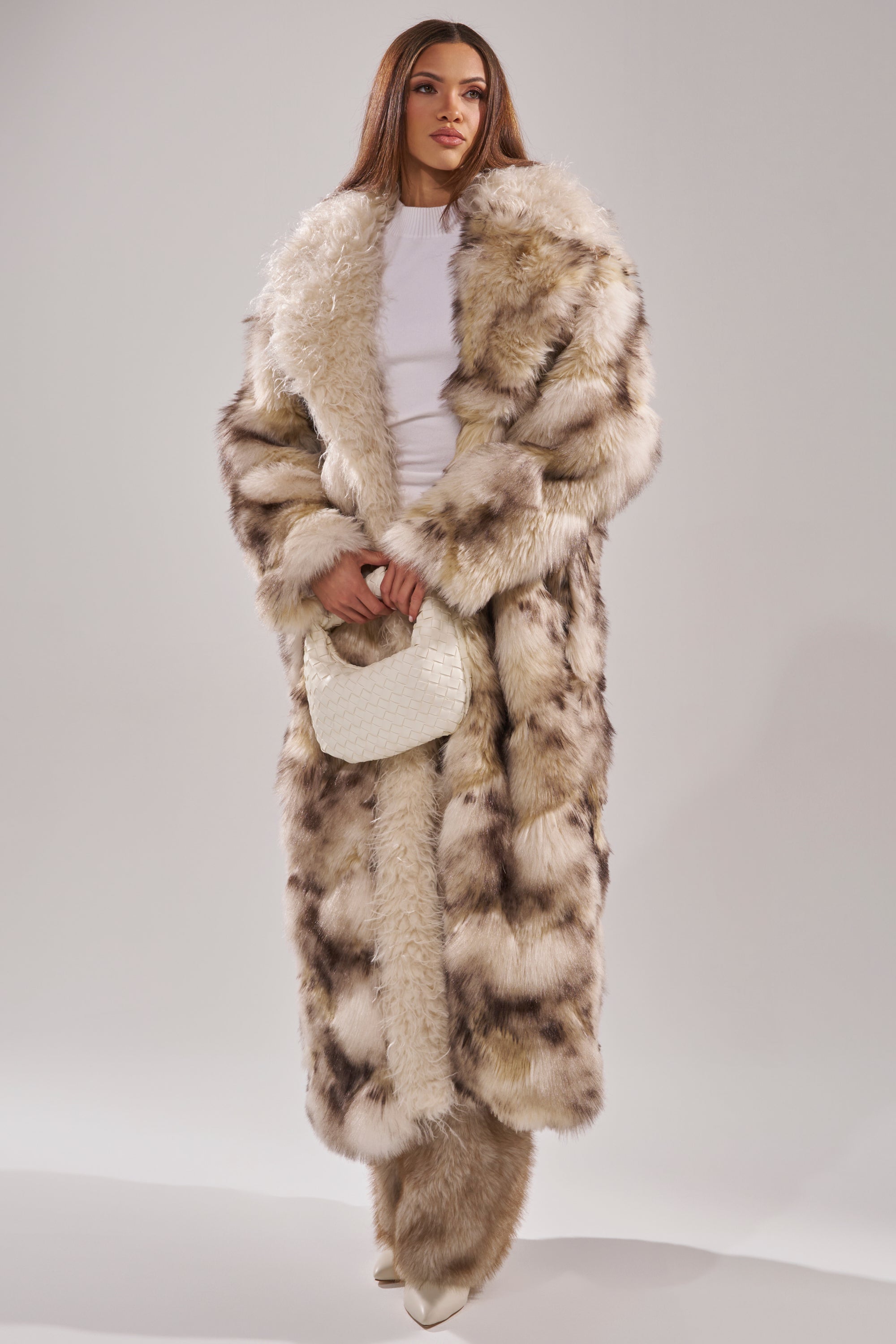 A woman models the PARK AVENUE PATCHWORK FAUX FUR, a full-length brown and white coat, over a white outfit with a small white handbag. She has long straight hair and neutral makeup, posing against a plain light background.