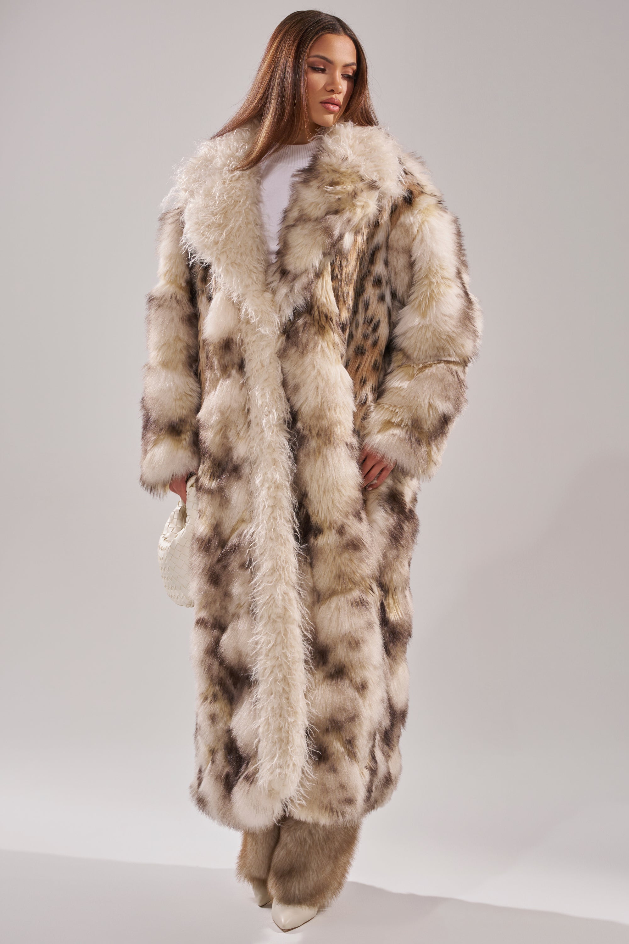 A woman stands against a plain light background, wearing the PARK AVENUE PATCHWORK FAUX FUR coat—full-length with cream, brown, and black spots—paired with white boots and a textured white handbag.