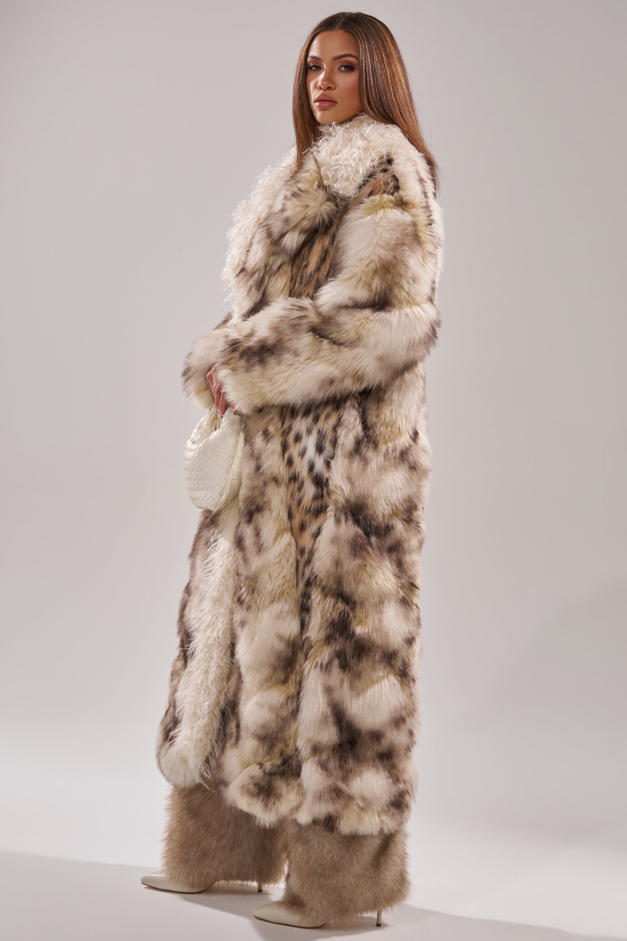 A woman stands confidently in the PARK AVENUE PATCHWORK FAUX FUR coat with matching furry pants, heeled boots, and a small white woven handbag against a plain light background.