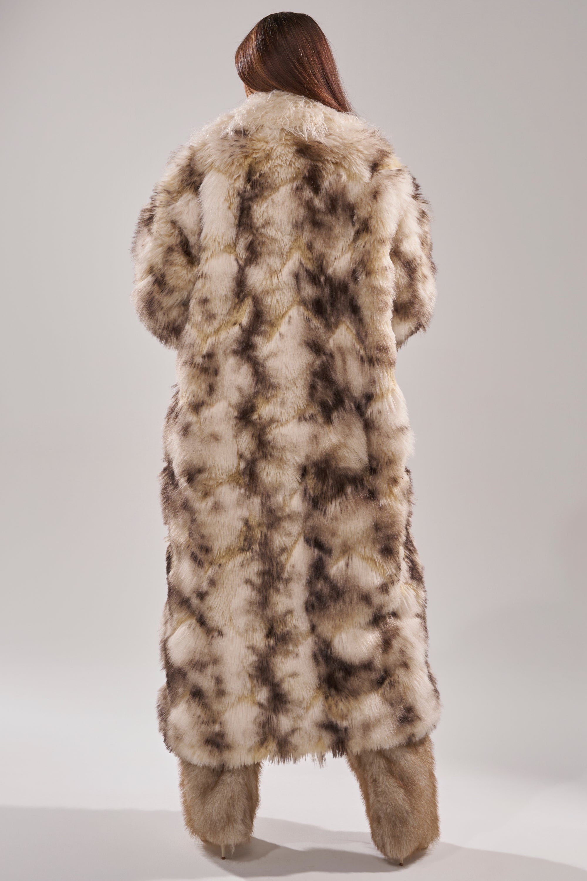 A person with long brown hair stands with their back to the camera, wearing the PARK AVENUE PATCHWORK FAUX FUR coat in cream and brown, paired with matching fur footwear against a plain light background.