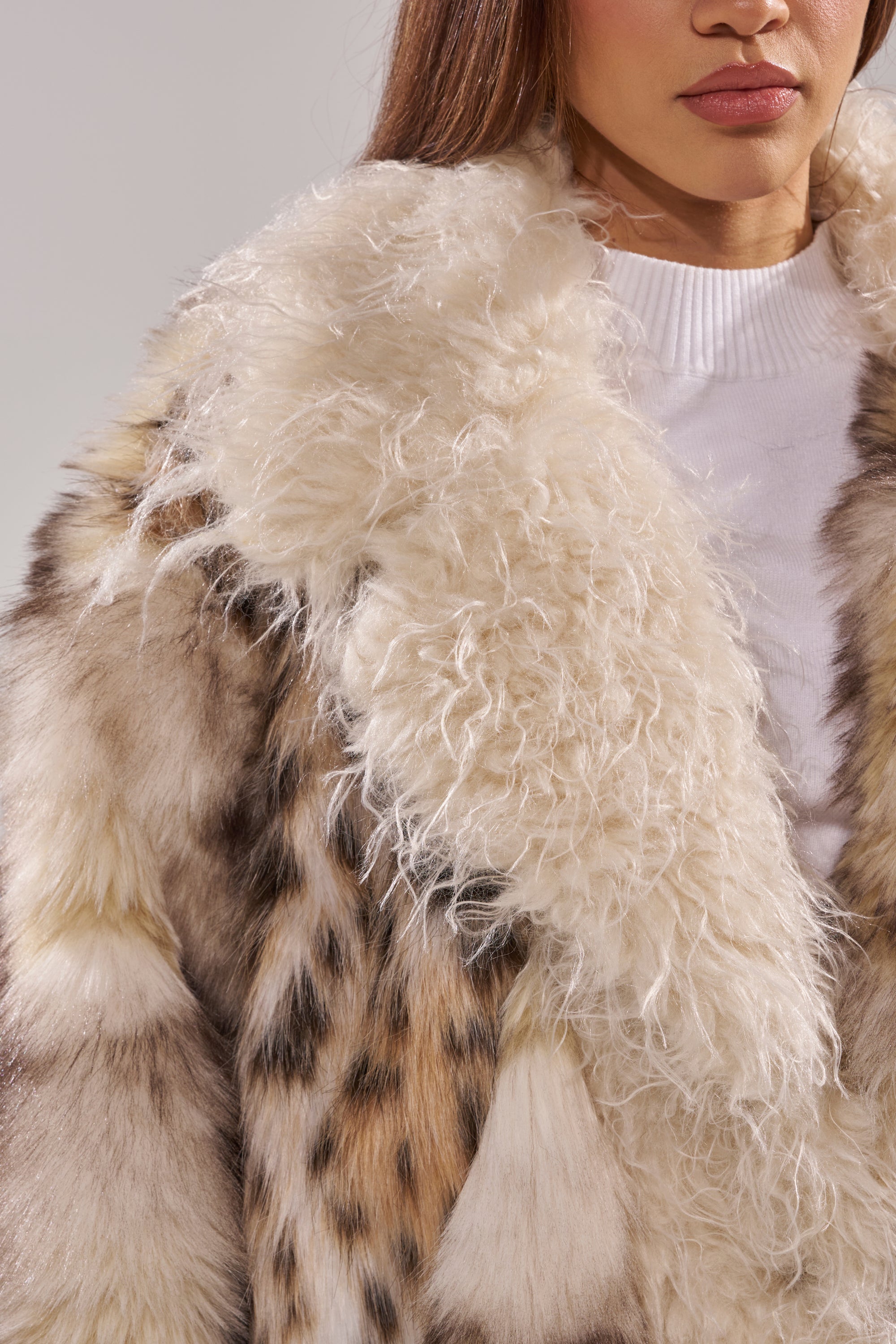 A woman models the PARK AVENUE PATCHWORK FAUX FUR coat, featuring a fluffy cream collar and mottled brown and black spots over a white sweater, with only her lower face and upper body visible.