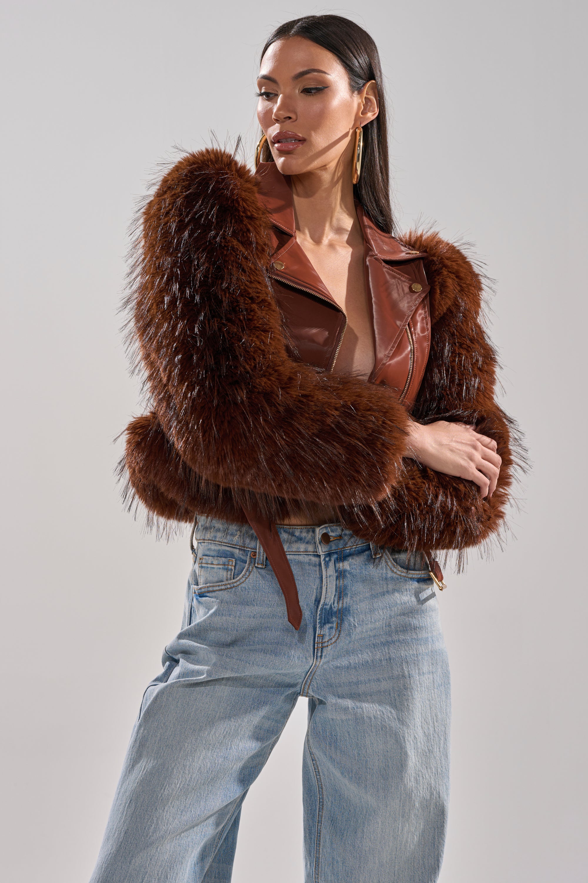 A woman models the SPEND IT ALL FUR TRIM MOTO brown faux leather jacket with light blue jeans, standing against a plain backdrop and looking sideways with her hands on her hips. 1