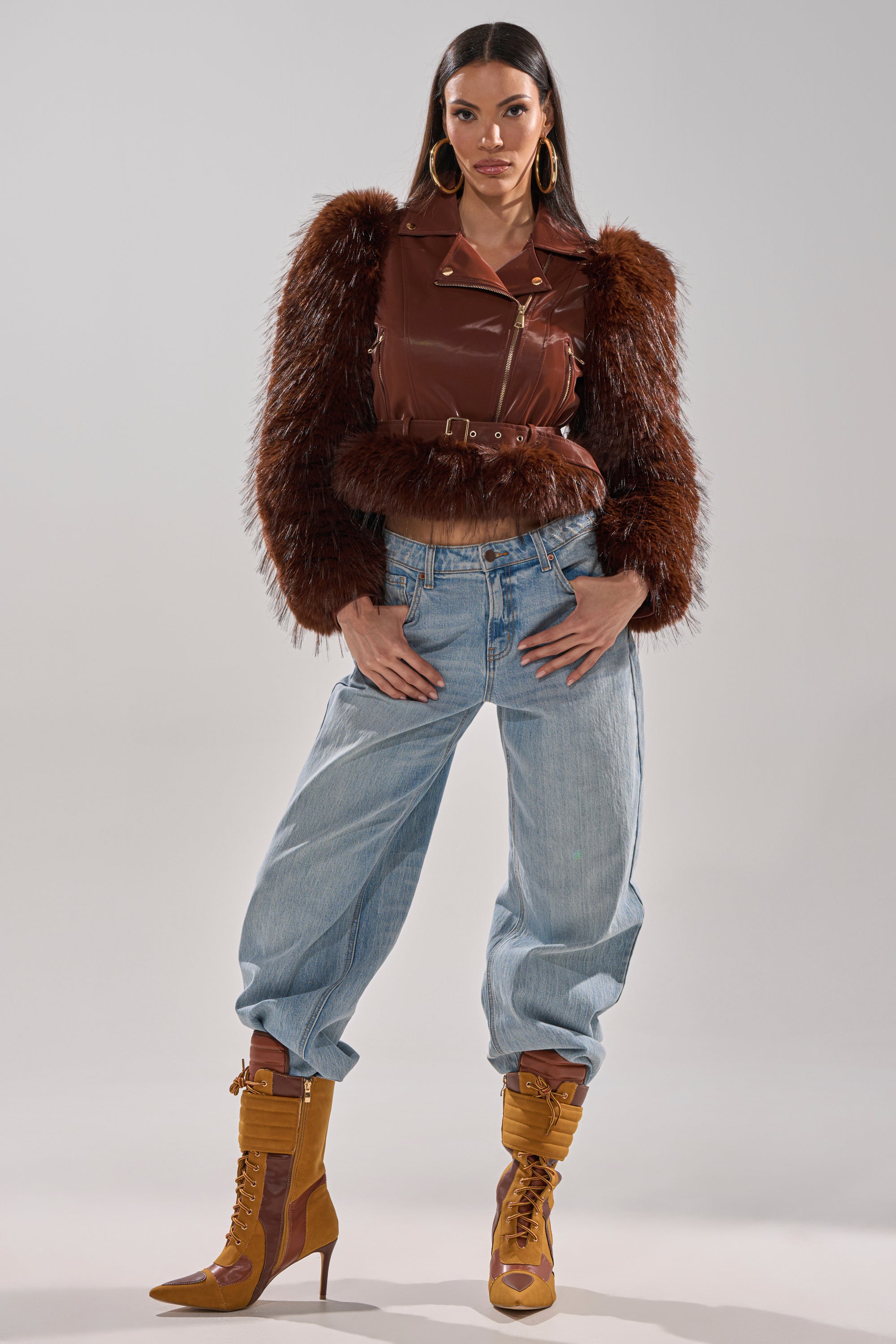 A woman stands confidently in the SPEND IT ALL FUR TRIM MOTO jacket with fur-trimmed sleeves, paired with light blue baggy jeans, mustard yellow high-heeled boots, and large hoop earrings against a plain light background. 9