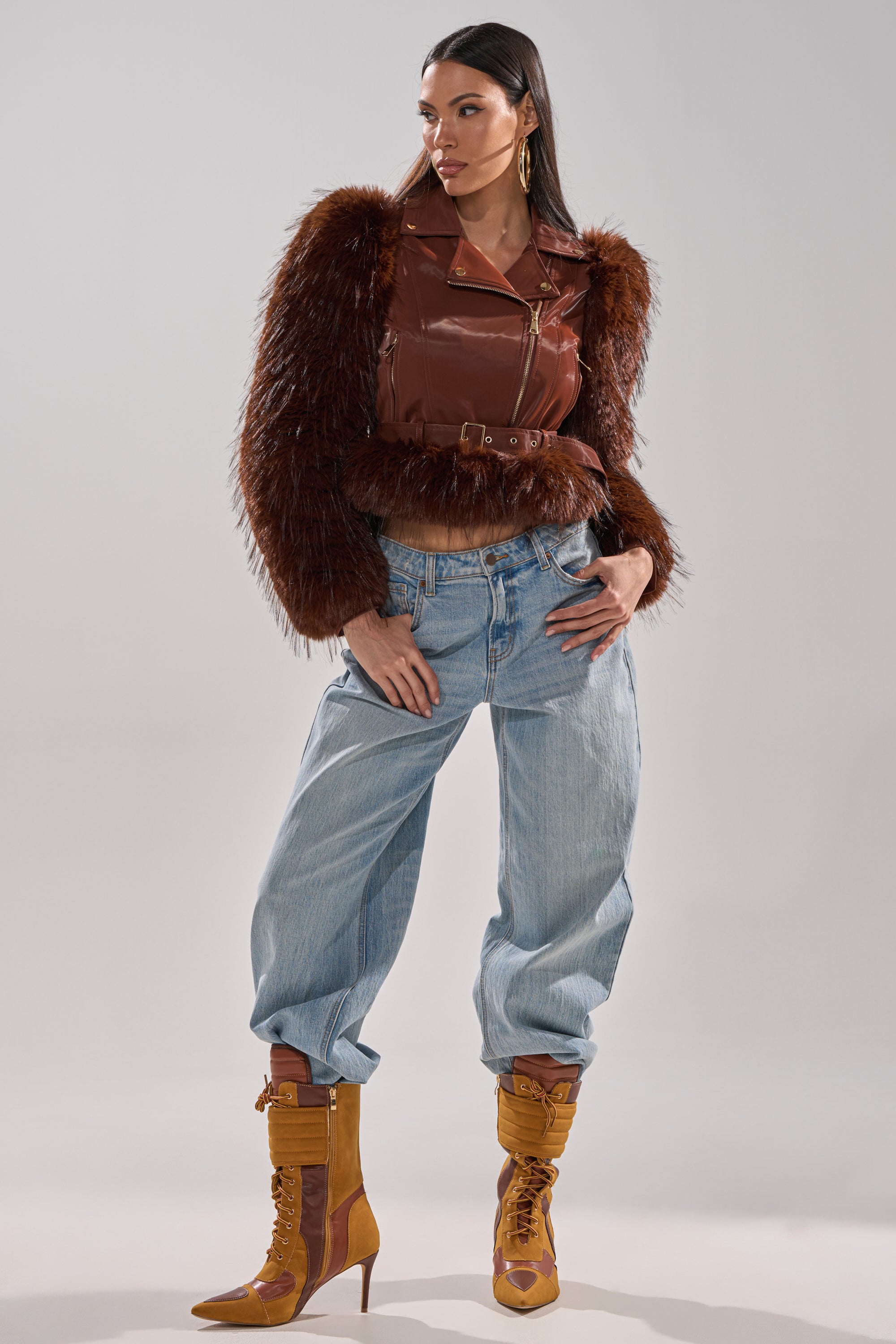 A woman with long dark hair and gold earrings stands against a neutral background, confidently wearing the SPEND IT ALL FUR TRIM MOTO jacket, light blue baggy jeans, and mustard-yellow lace-up heeled boots with faux fur sleeves. 10