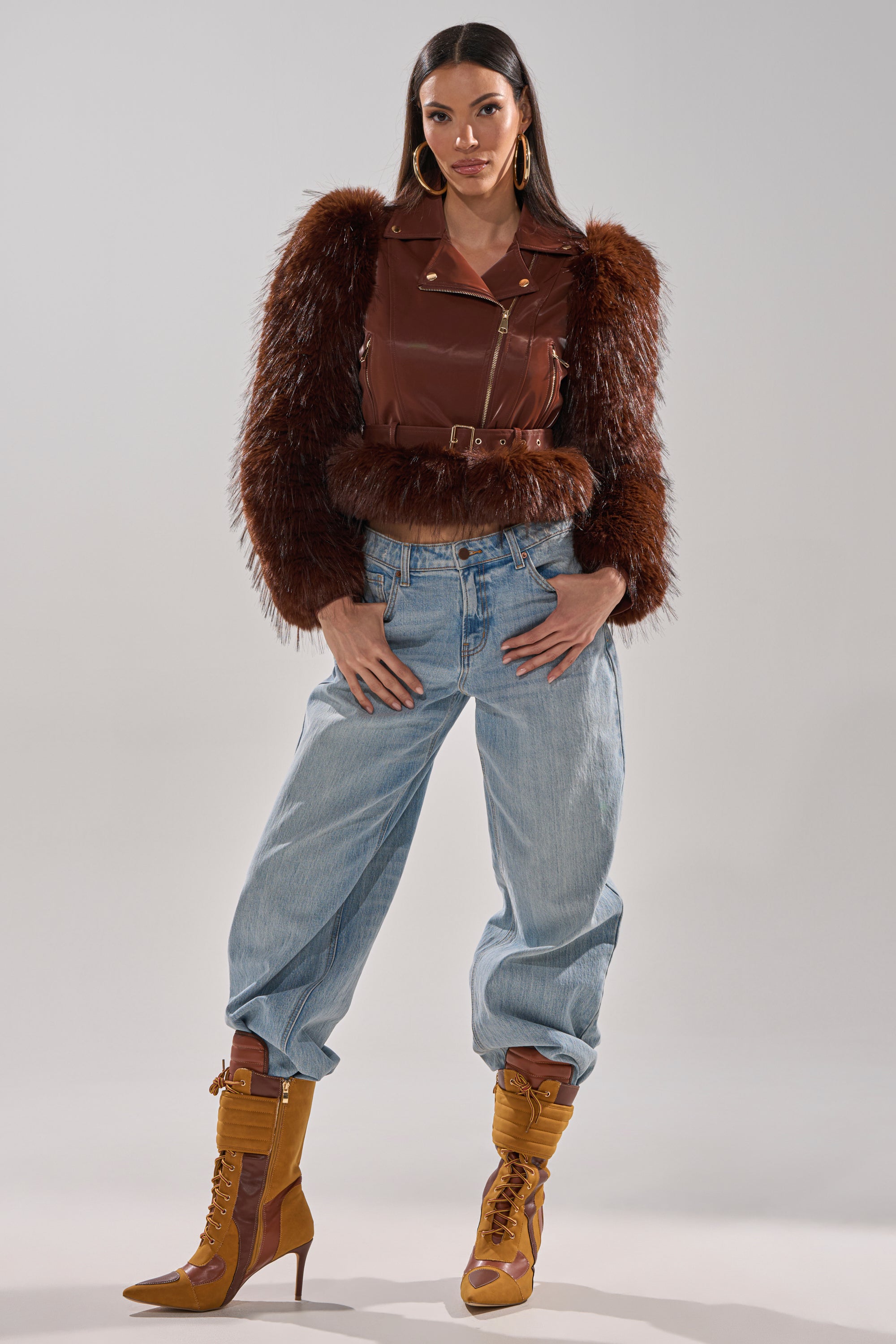 A woman with long dark hair and hoop earrings poses confidently against a plain background, wearing the SPEND IT ALL FUR TRIM MOTO— a brown faux leather moto jacket with faux fur sleeves—light blue baggy jeans, and tan heeled boots. 11