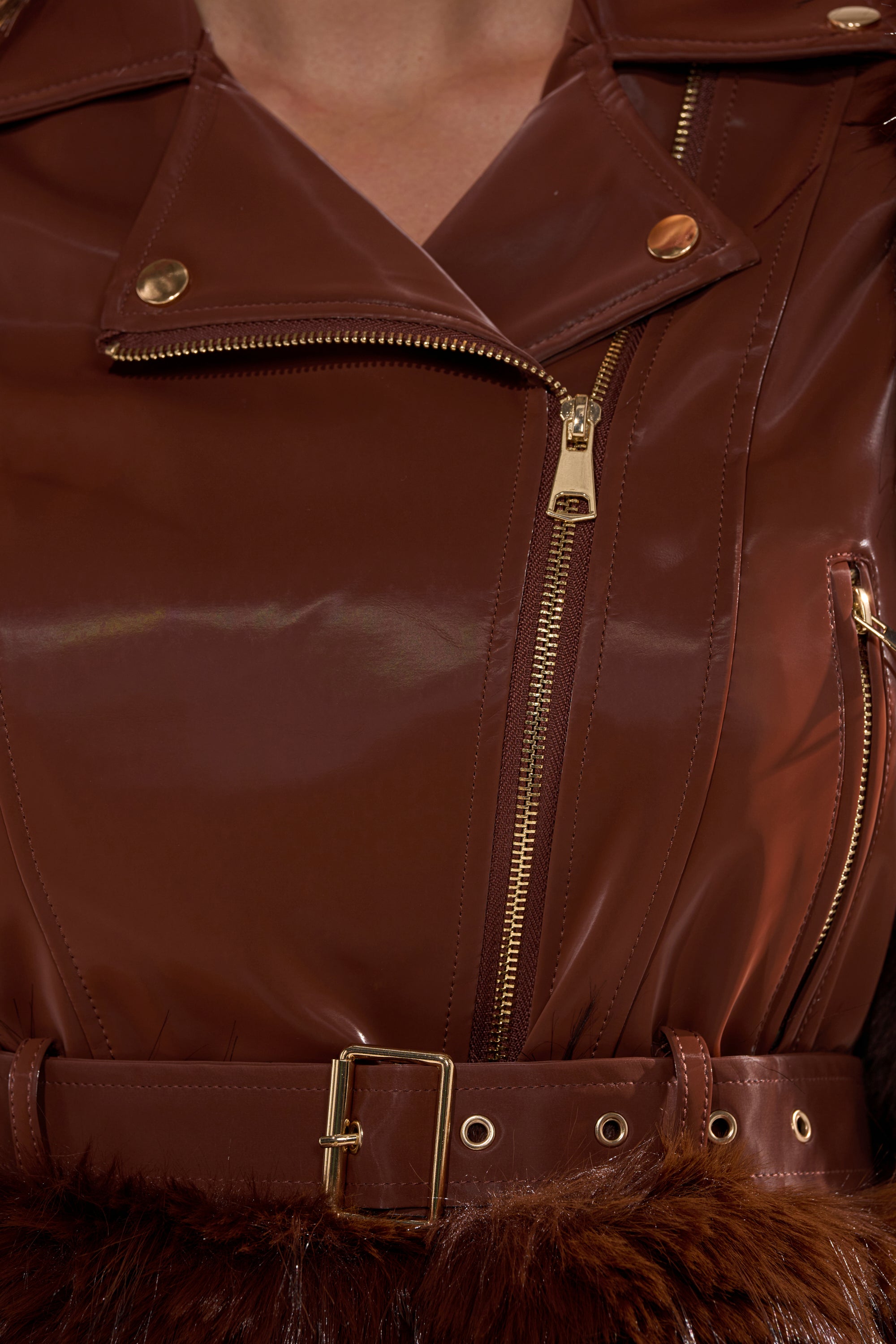 Close-up of the SPEND IT ALL FUR TRIM MOTO jacket—shiny brown faux leather, gold zippers, snap buttons, and a matching belt with gold buckle. The lower edge features soft brown fur trim for a luxe finish. 12
