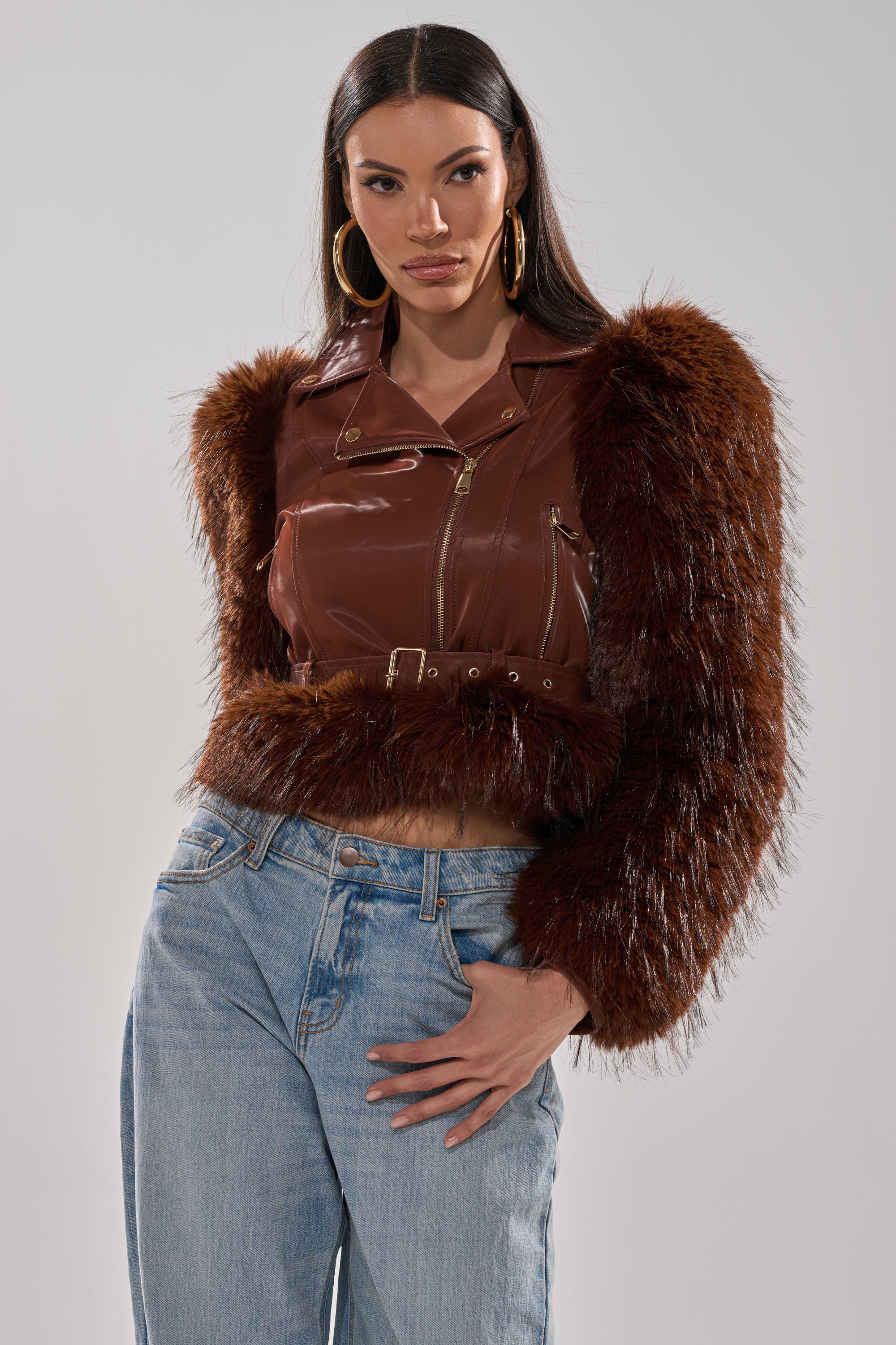 A woman models the SPEND IT ALL FUR TRIM MOTO, a brown faux leather jacket with faux fur sleeves, gold zippers, and a belt, styled with light blue jeans and hoop earrings, against a plain background. 0