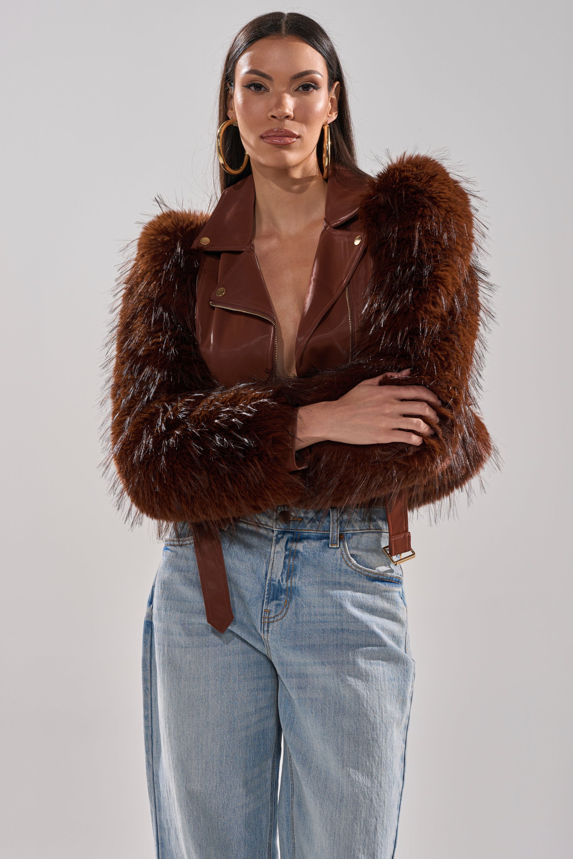 A woman in the SPEND IT ALL FUR TRIM MOTO jacket with brown faux fur sleeves, gold buttons, and light blue high-waisted jeans stands with her arms crossed against a plain light gray background. 2