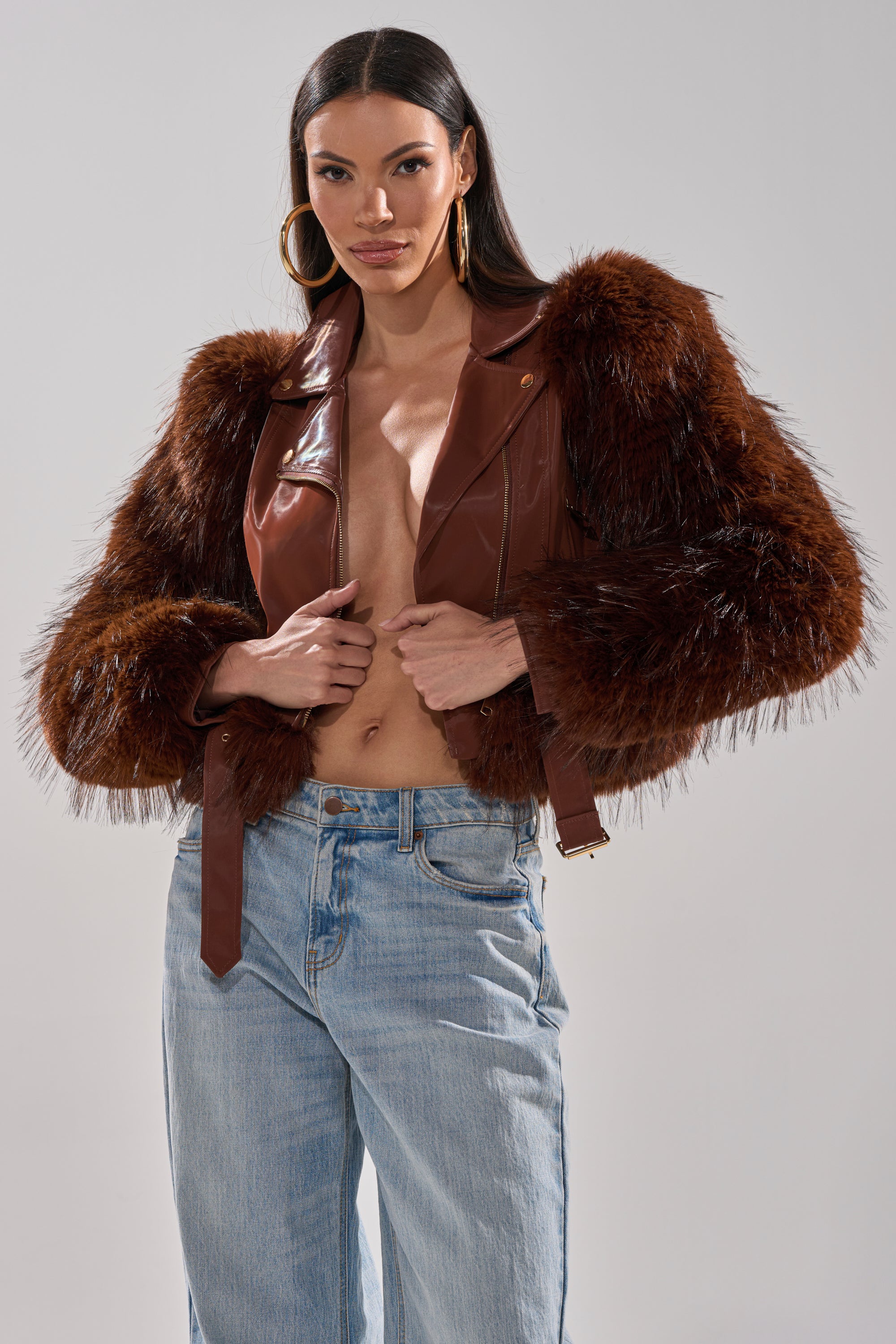 A woman models the SPEND IT ALL FUR TRIM MOTO— a brown faux leather moto jacket with faux fur sleeves—paired with light blue jeans. She has long dark hair, large hoop earrings, and poses with her hands near the jacket’s front. 3