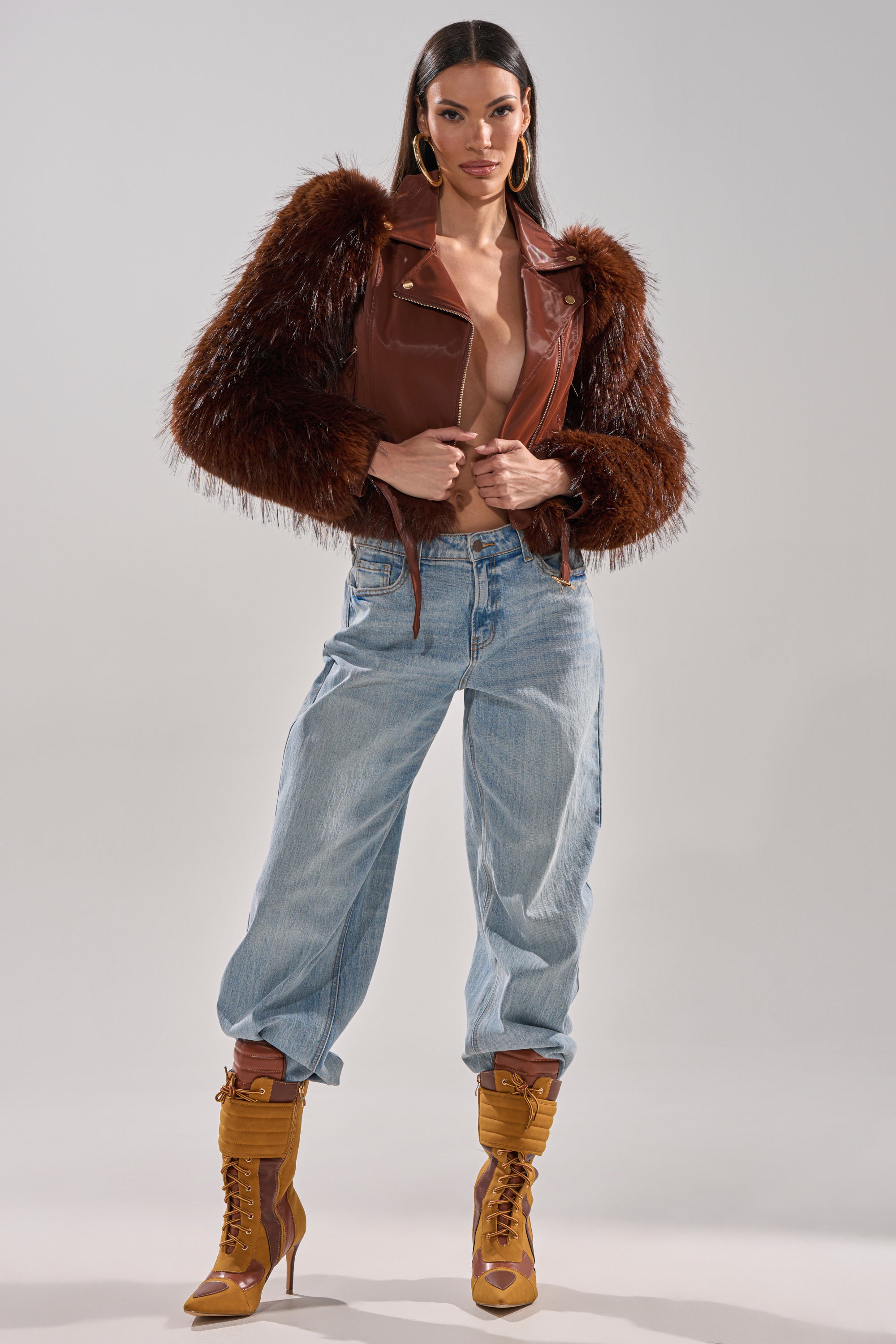 A woman exudes confidence in the SPEND IT ALL FUR TRIM MOTO jacket with furry sleeves, styled with loose light blue jeans, tan high-heeled boots, gold hoop earrings, and long straight hair against a plain gray backdrop. 8