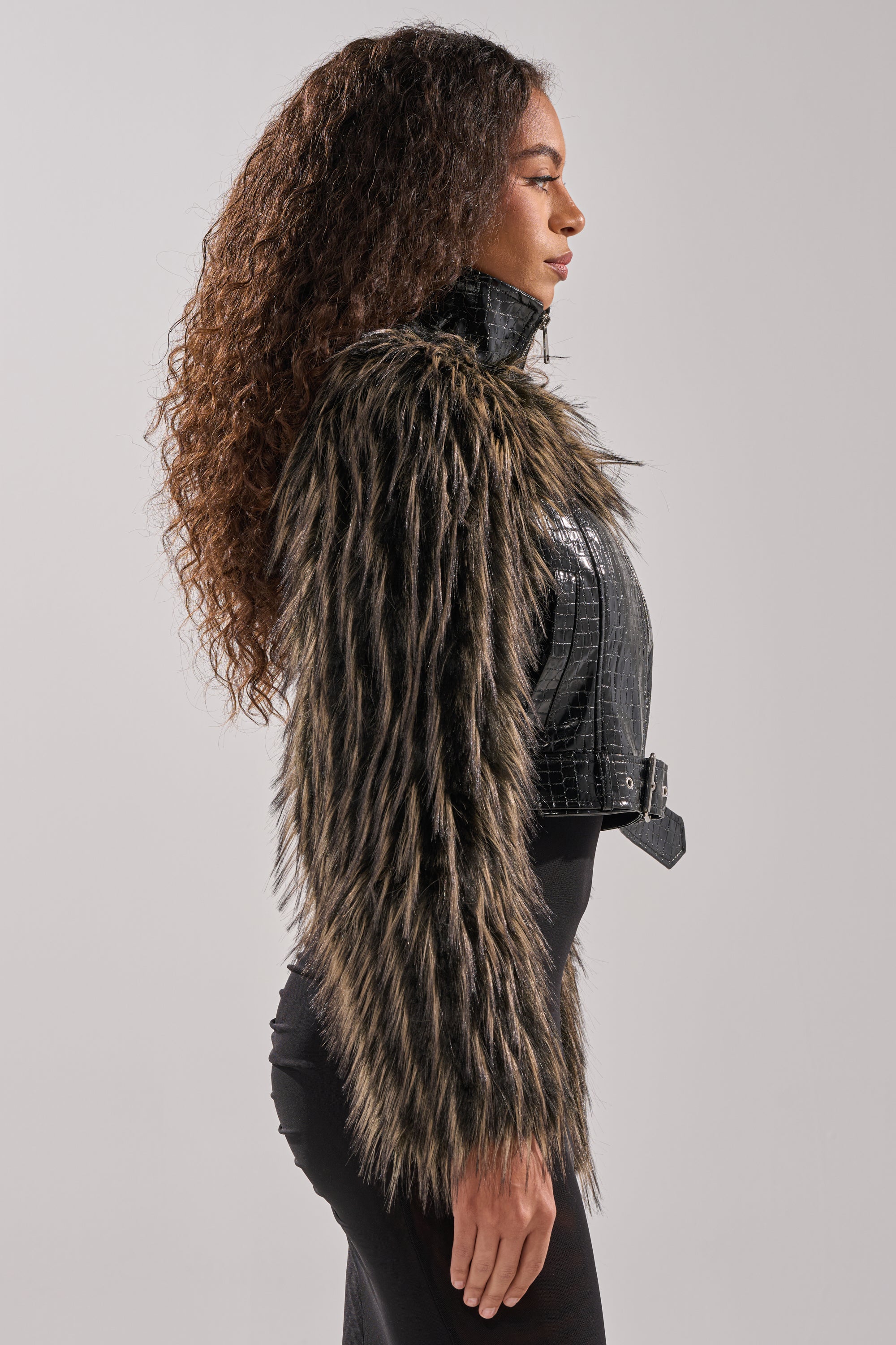 A woman in profile wears the LIGHT IT UP FUR TRIM MOTO, a cropped jacket with long black faux fur trim and a textured pattern, paired with a fitted black skirt against a plain light background. 5