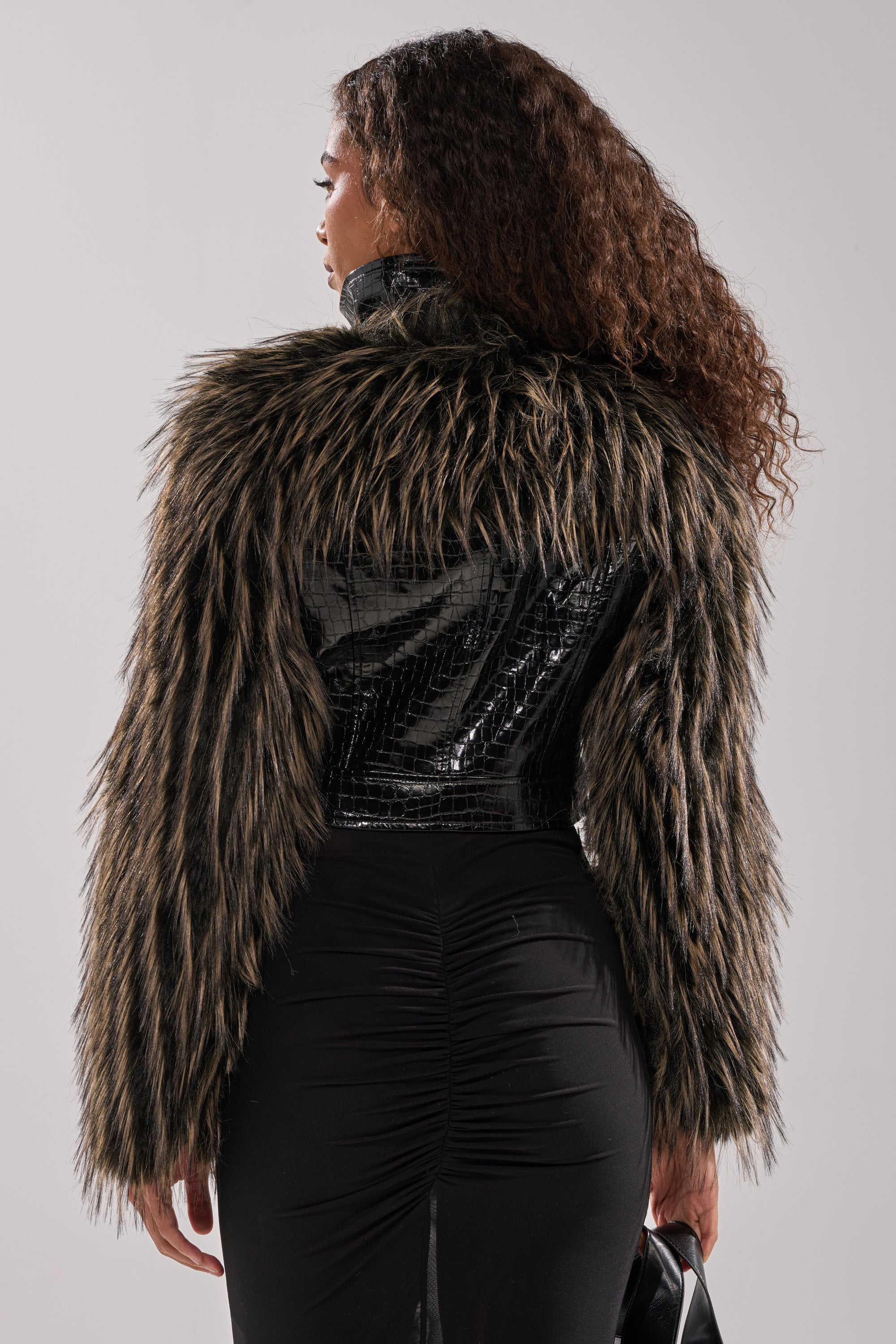 A person with long curly hair, seen from behind, wears the LIGHT IT UP FUR TRIM MOTO—a black faux crocodile leather jacket with shaggy faux fur sleeves and collar—paired with a fitted black skirt. 6
